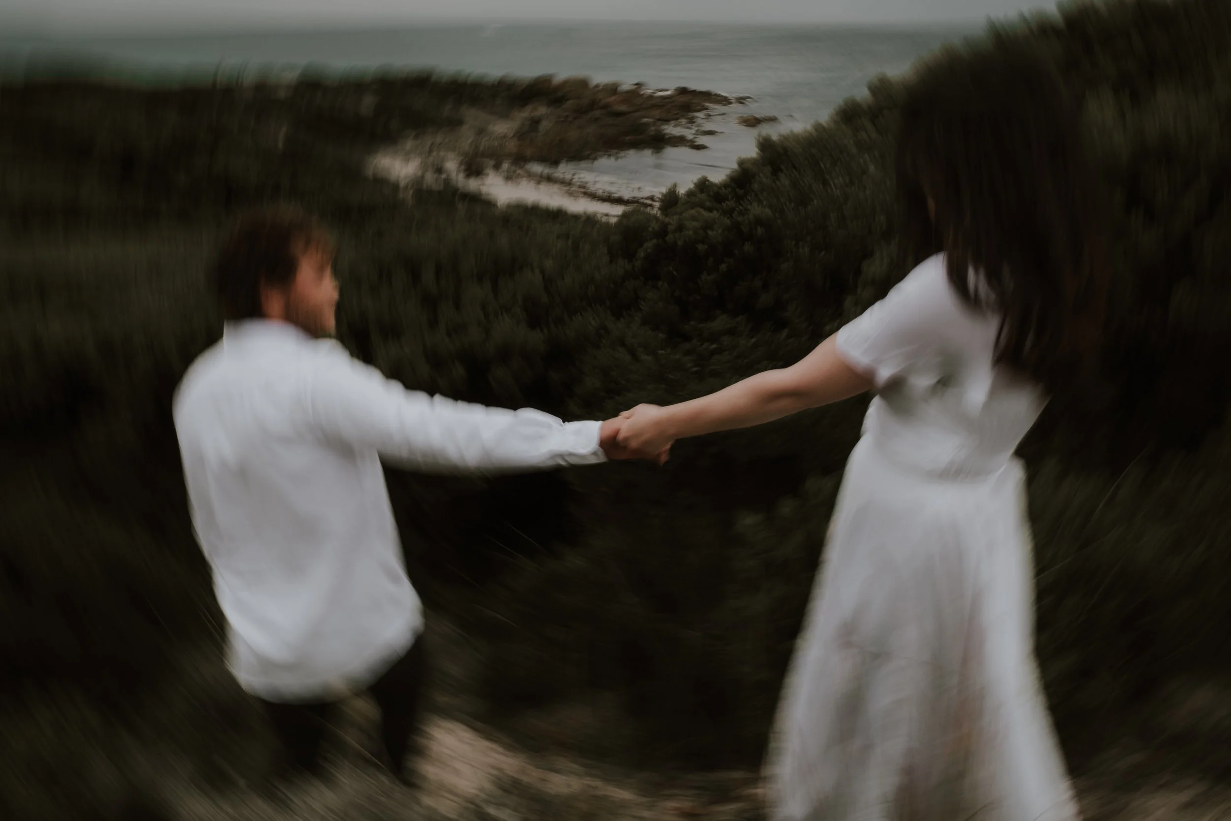 A blurry photo of a man and woman holding hands, walking outdoors near the coast, with the sea and rocks in the background.