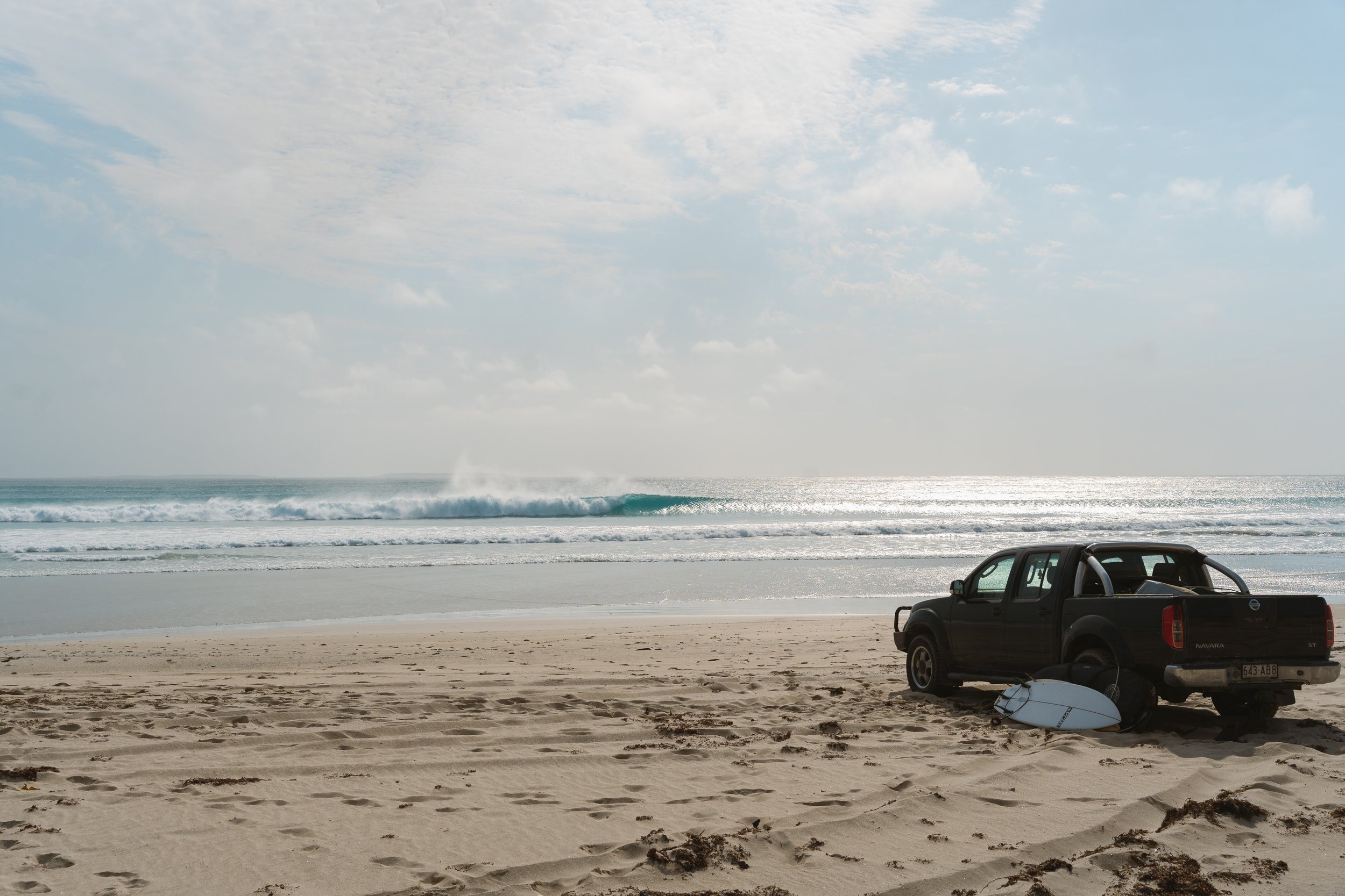 A black pickup truck parked on a sandy beach near the ocean, with a white surfboard lying on the sand nearby. Waves are crashing in the ocean under a partly cloudy sky.