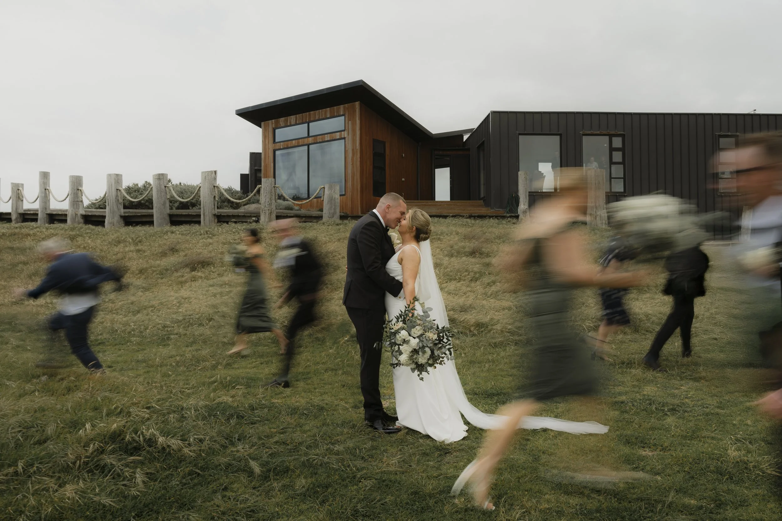 A bride and groom smiling and touching foreheads outdoors with a modern house in the background. The scene includes blurred motion of people running or walking past.