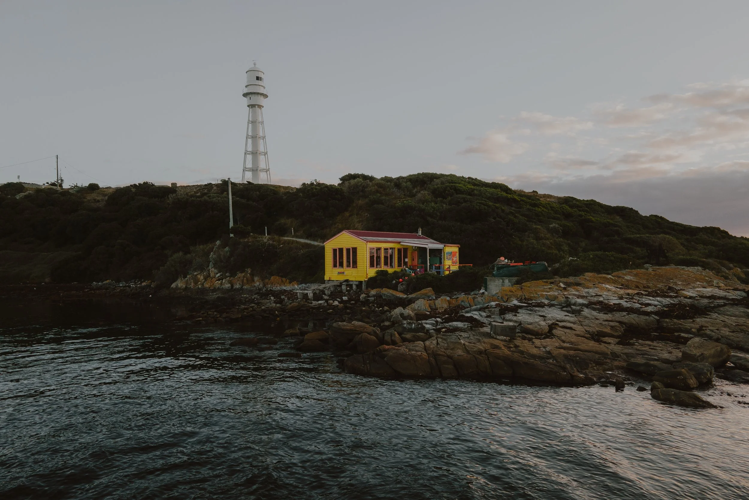 A yellow house with a red roof situated on rocky terrain at the water's edge, with a large white communication tower on a hill and a partly cloudy sky in the background.