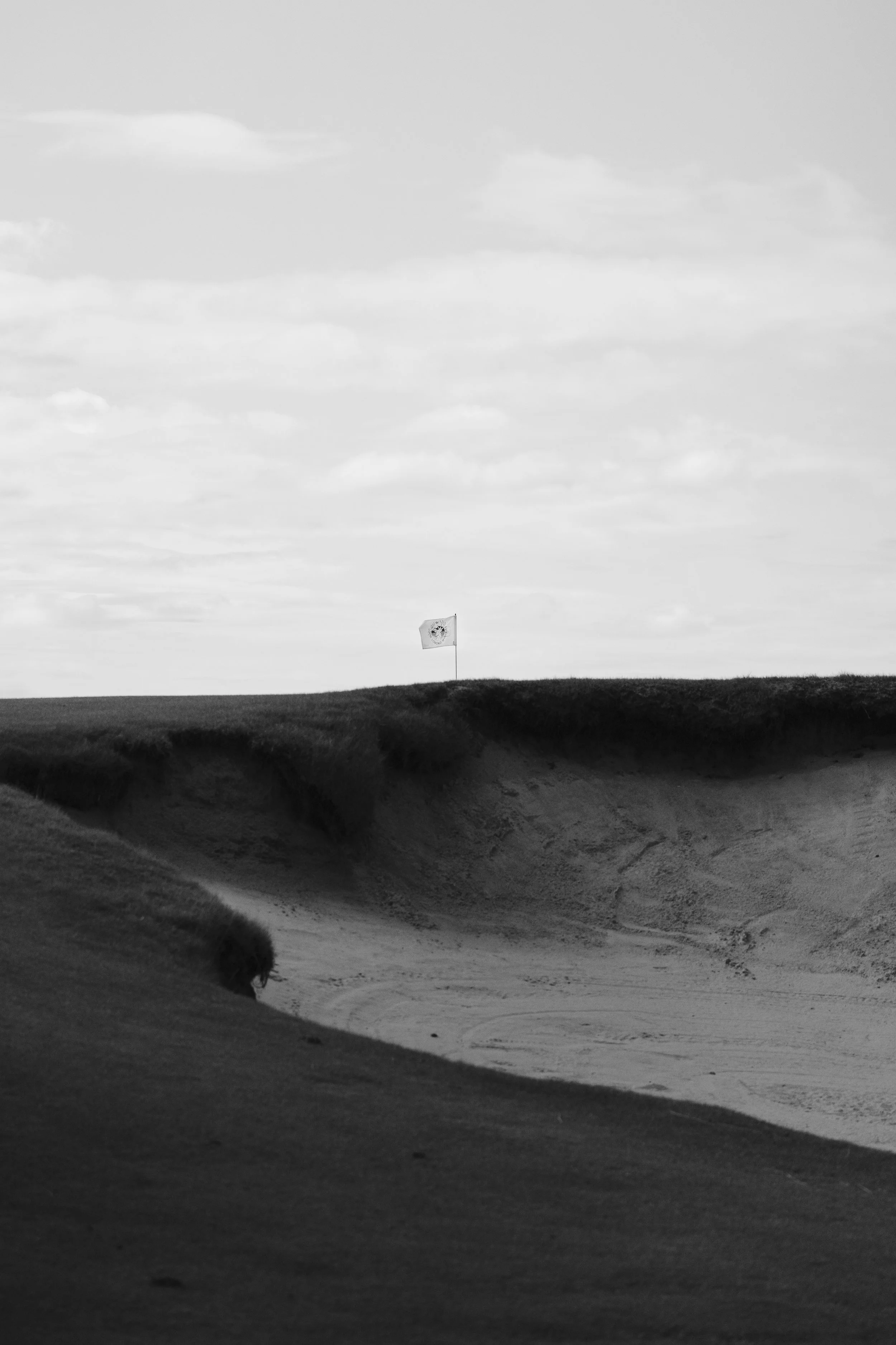 A black and white photo of a grassy landscape with sand dunes in the foreground and a lone flag on a pole in the distance under a cloudy sky.