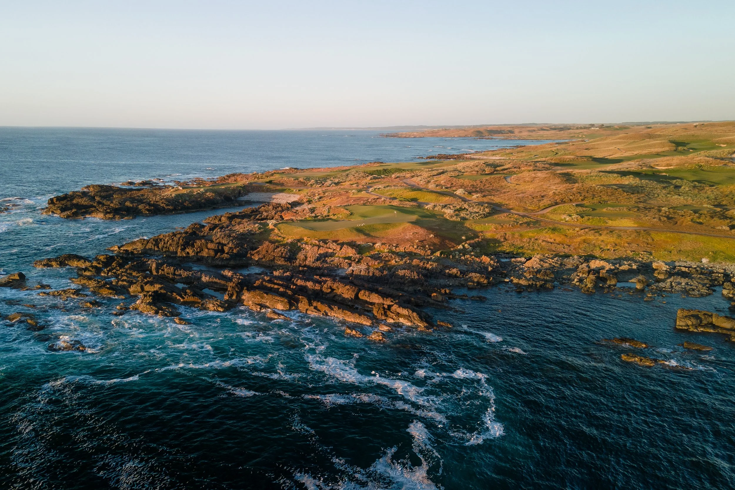 Aerial view of a rugged coastline with rocky cliffs and patches of grassy terrain, meeting the ocean's waves under a clear sky.