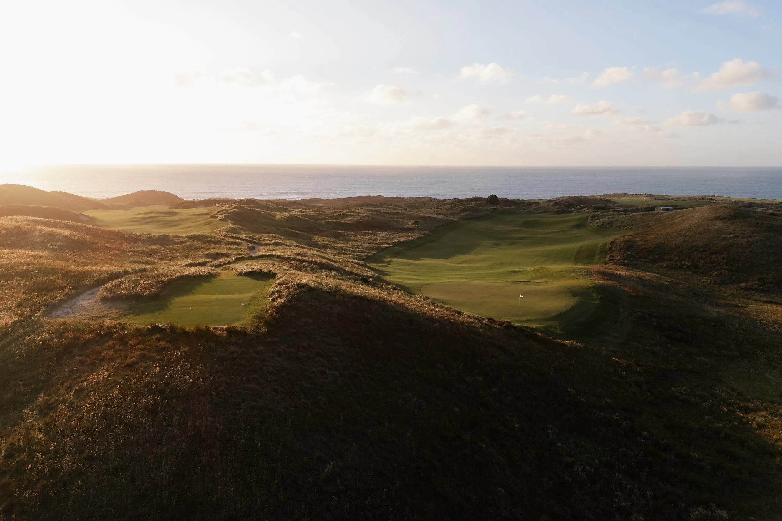A scenic view of a golf course with rolling hills near the ocean under a partly cloudy sky during sunset.