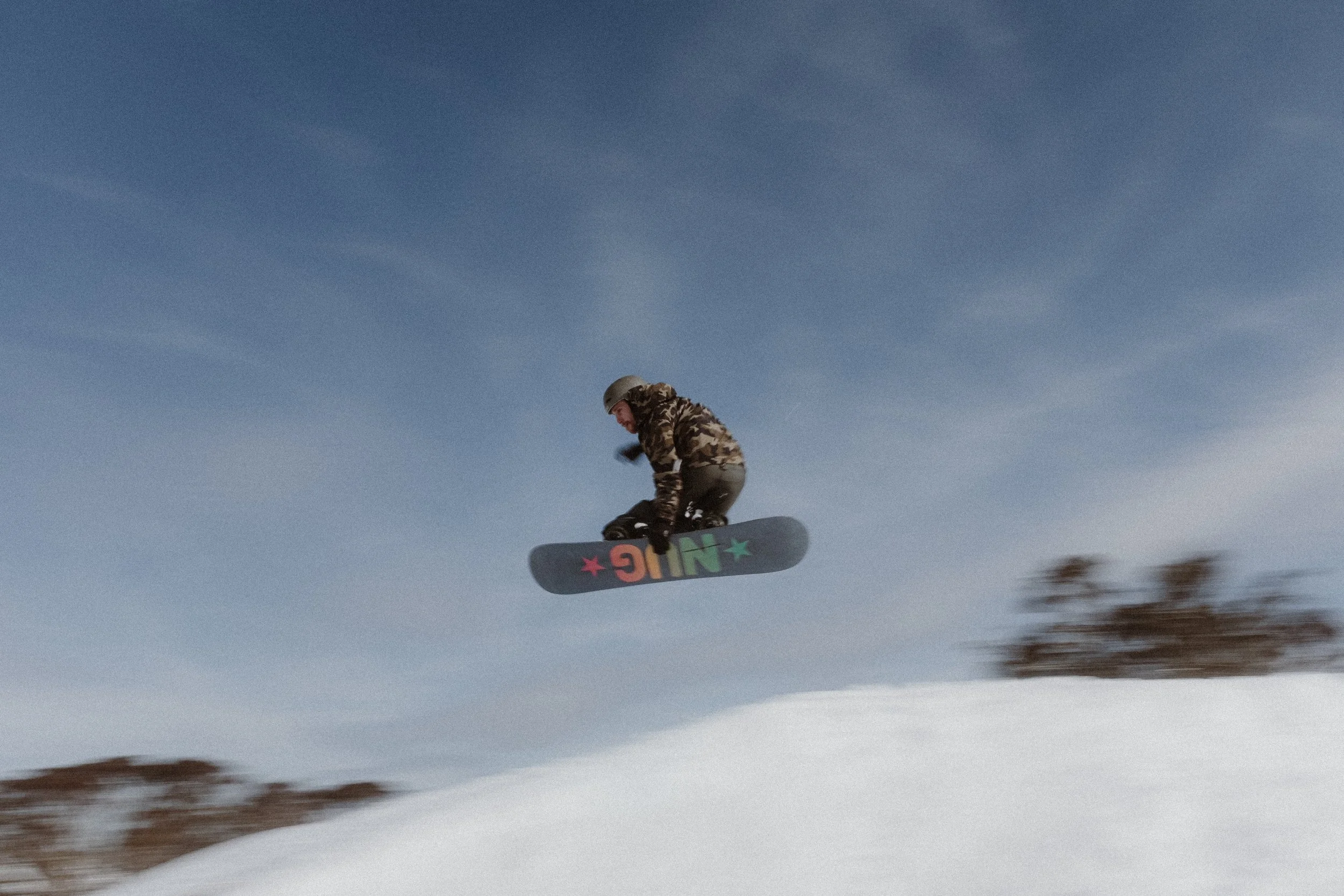 A person dressed in camouflage clothing and wearing a helmet is riding a skateboard in mid-air against a clear blue sky.
