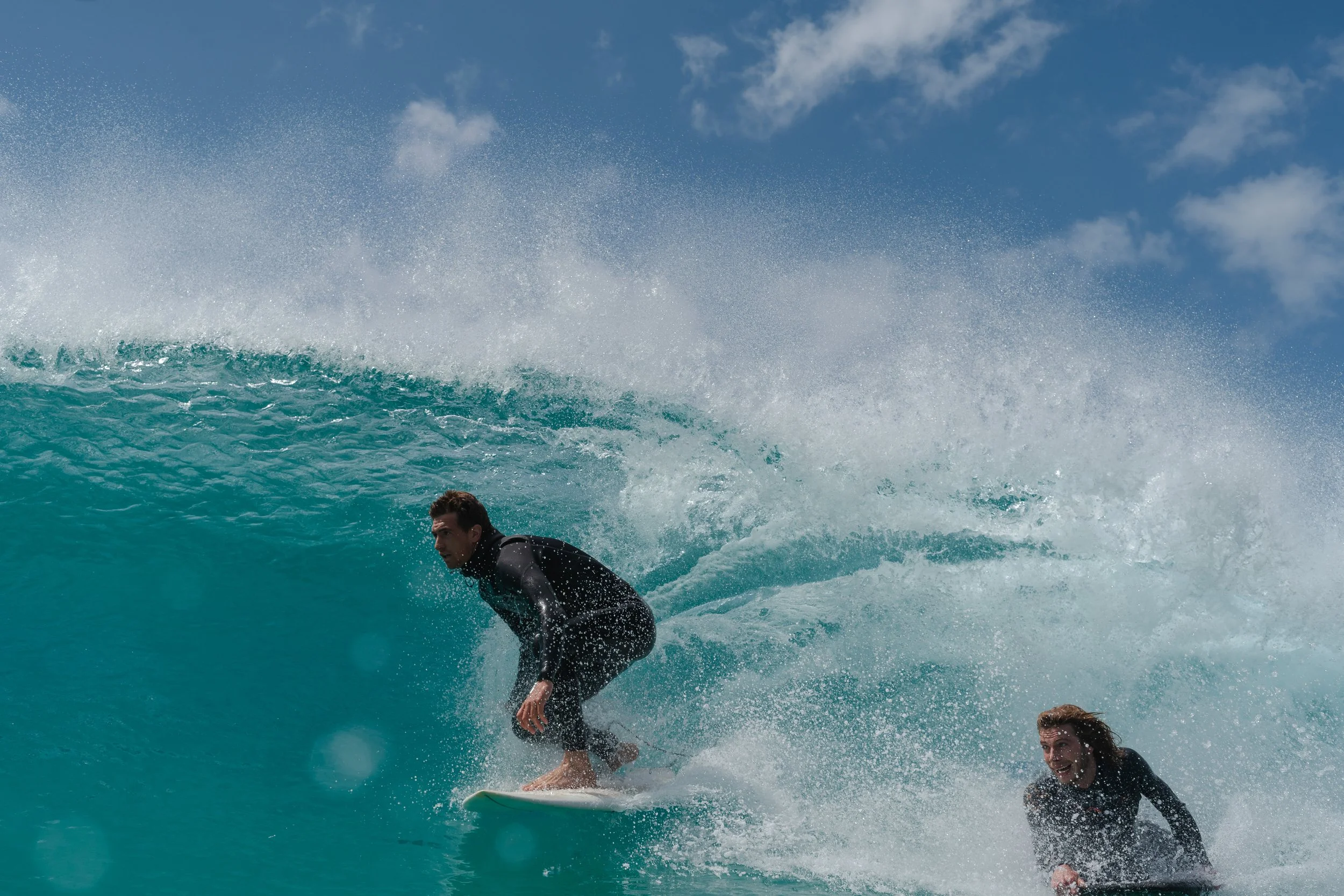 Two surfers riding a wave in the ocean with clear blue sky and some clouds.