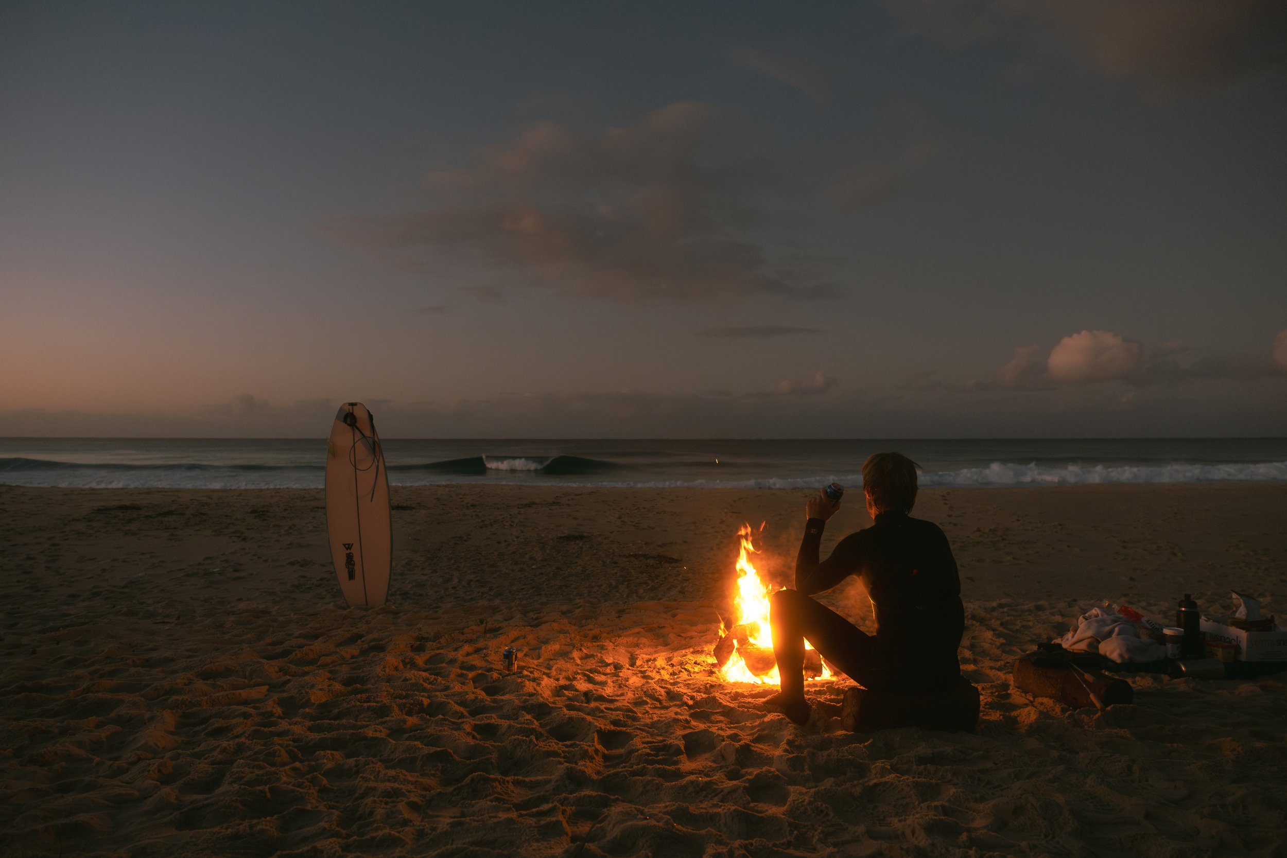 Person sitting by a campfire on a sandy beach at dusk, with surfboards standing upright in the sand and clouds in the sky.