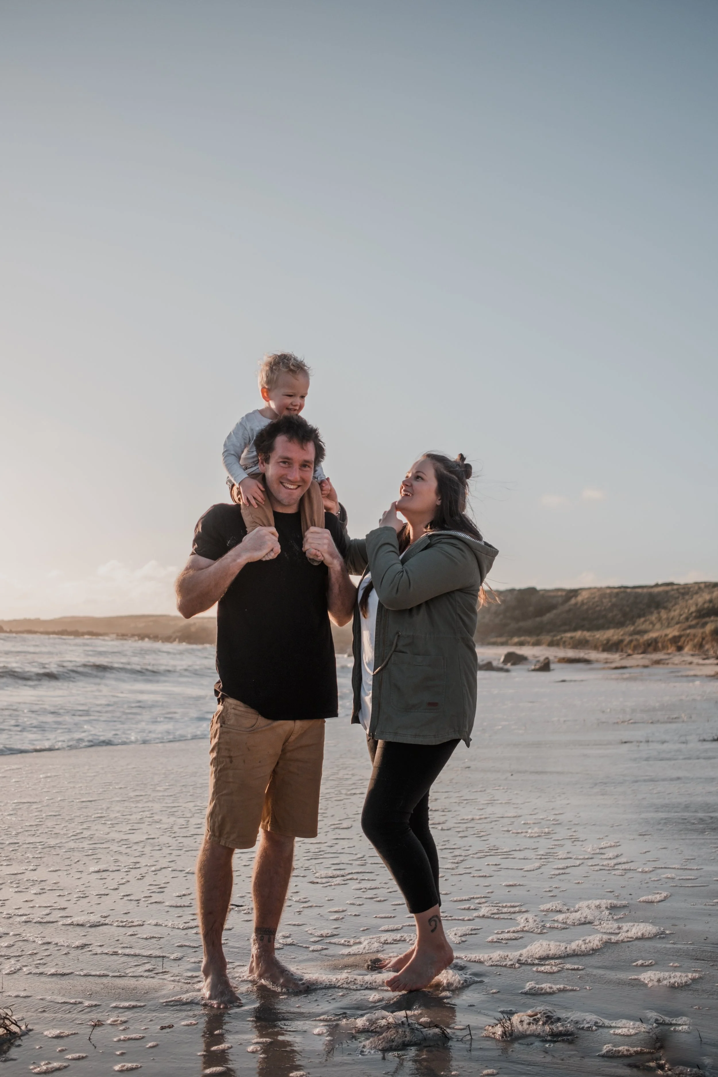 A family of three with light skin at the beach during sunset. The father is carrying a young child on his shoulders, and the mother is standing next to them, smiling and looking at the child. They are barefoot, standing on wet sand with gentle waves 