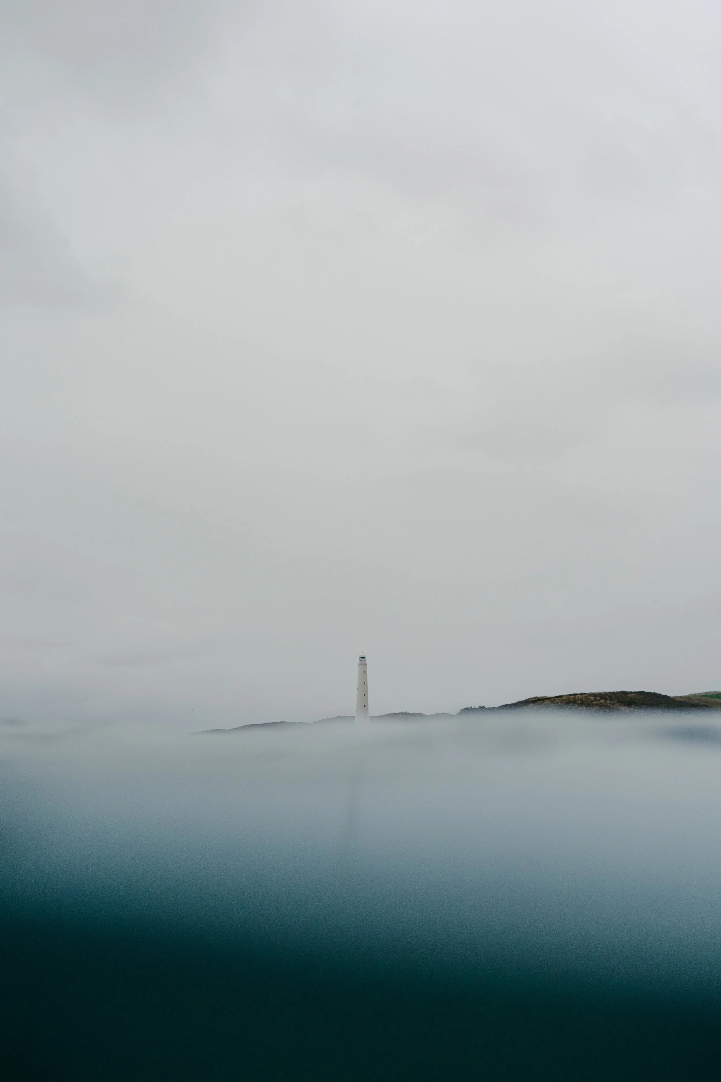 A lighthouse on a small hill surrounded by low clouds or fog, with cloudy skies above.