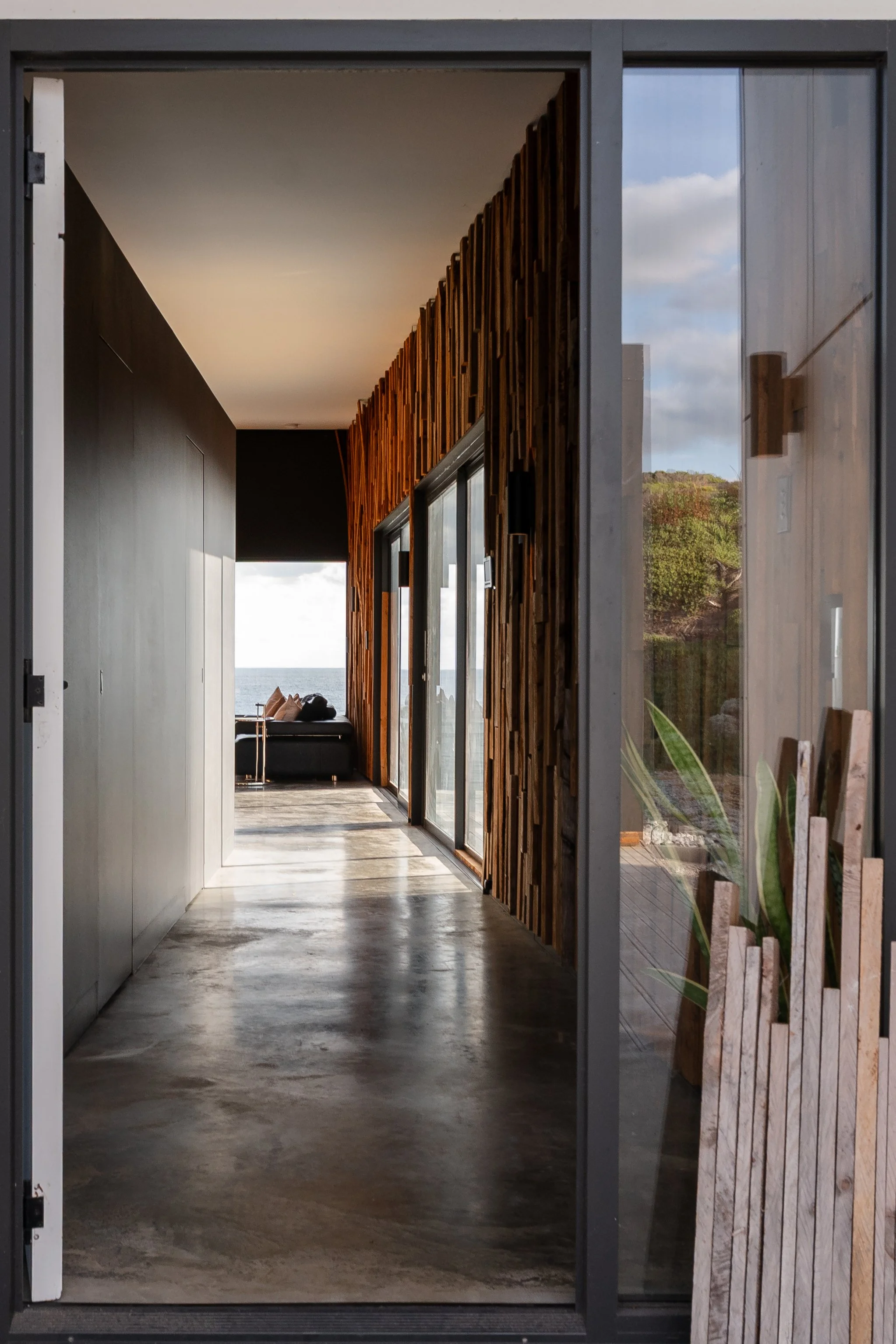 View through a glass door into a modern hallway with wooden wall panels on the right, leading to a living area with a black sofa and view of the ocean outside.