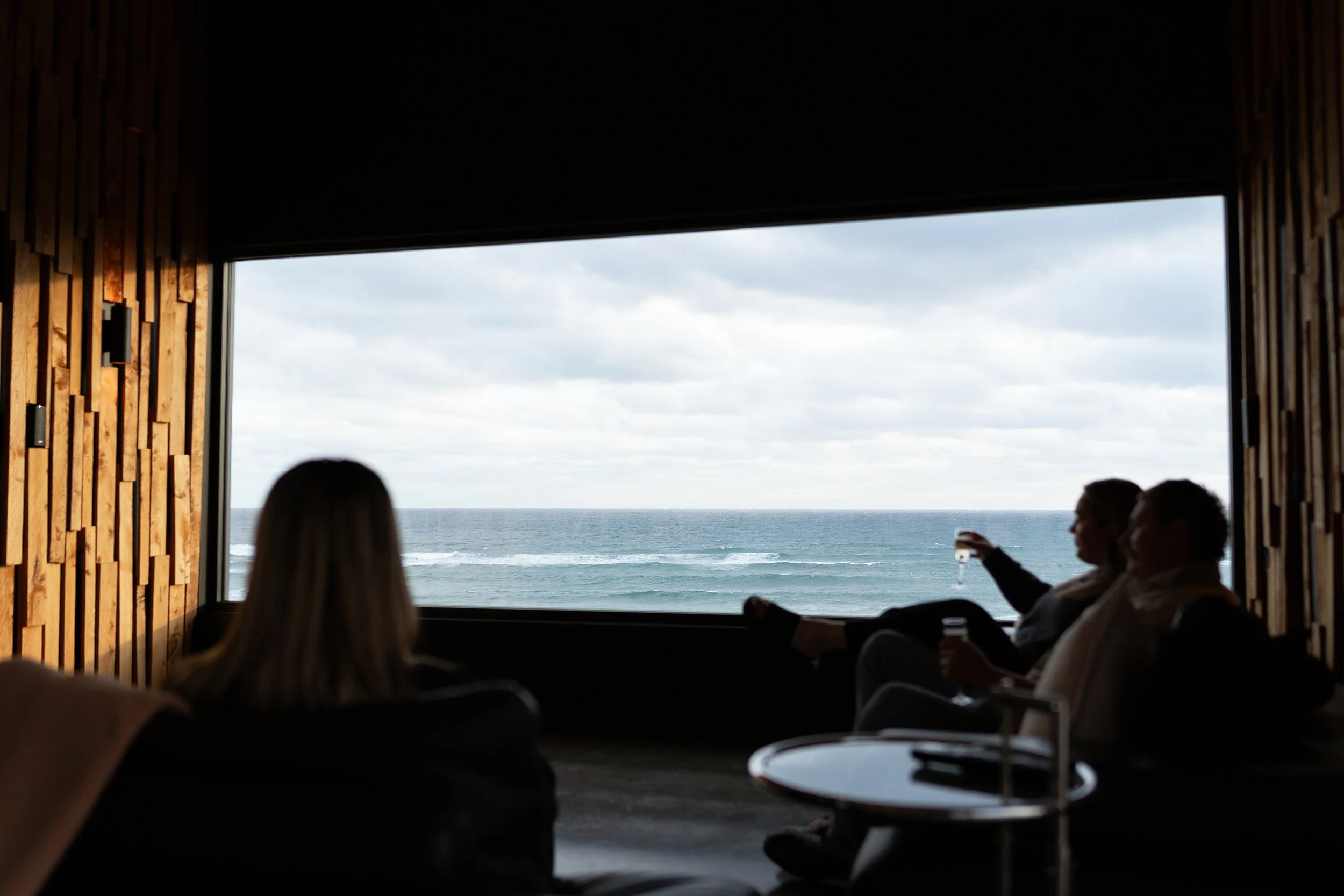 People sitting indoors near large window overlooking the ocean with cloudy sky.
