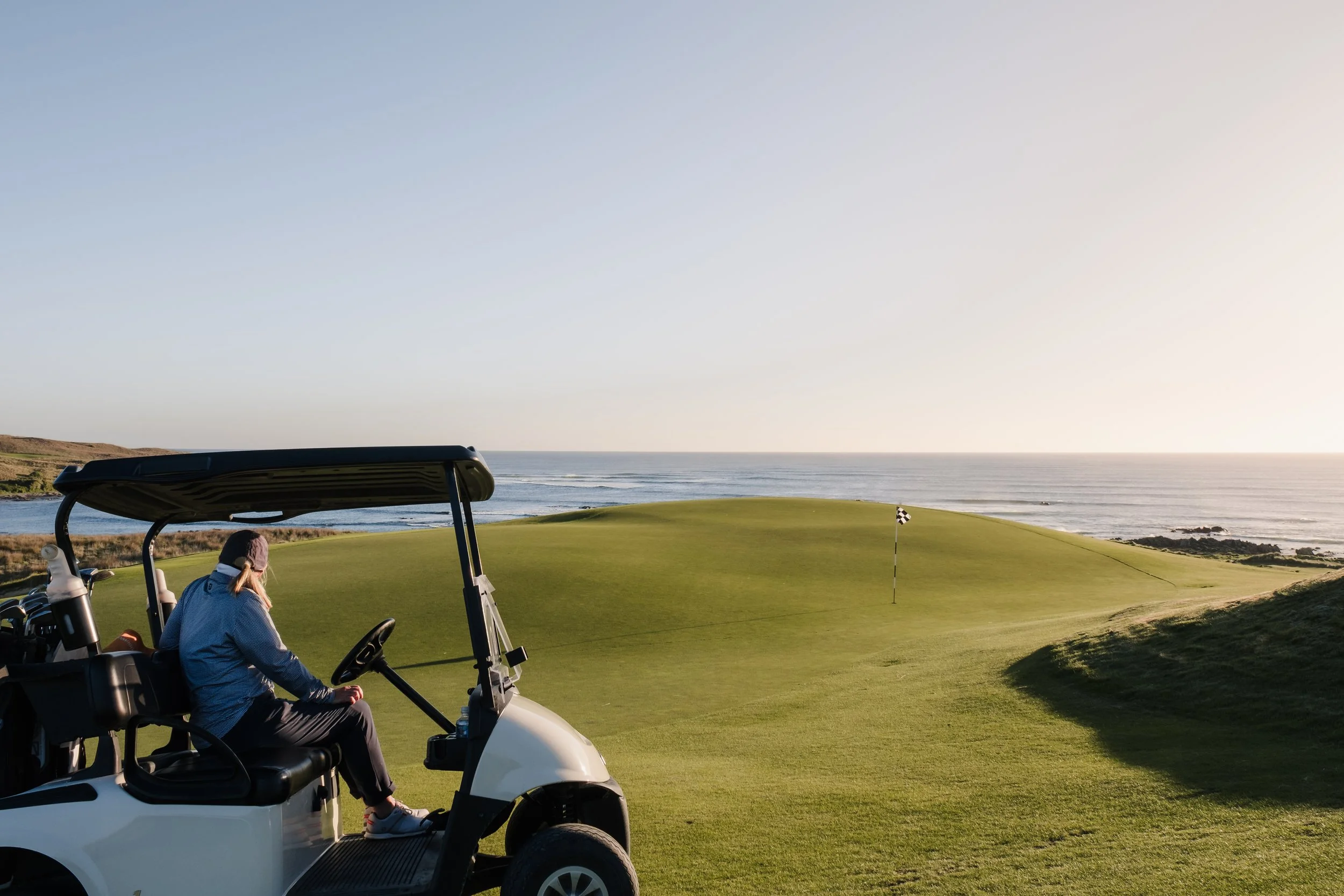 A woman sitting in a golf cart on a golf course, with a view of a green, a flag, and the ocean in the background during sunset.