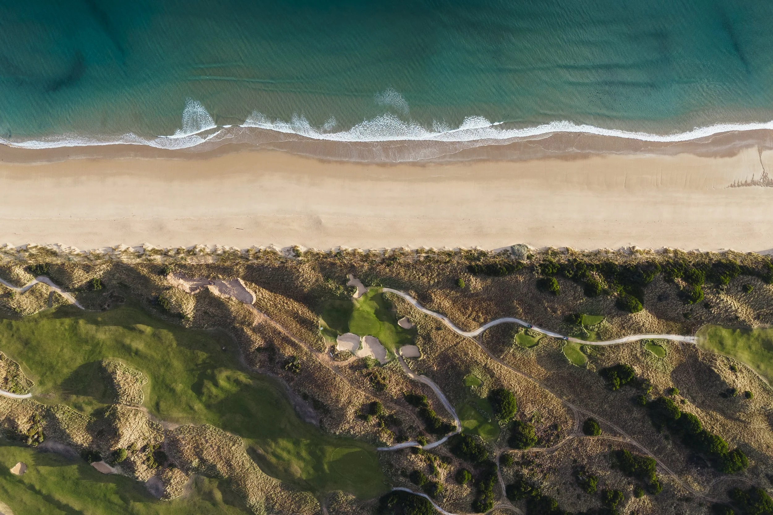 An aerial view of a beach with turquoise water and white waves, adjacent to a sandy shoreline with patches of vegetation and winding paths.