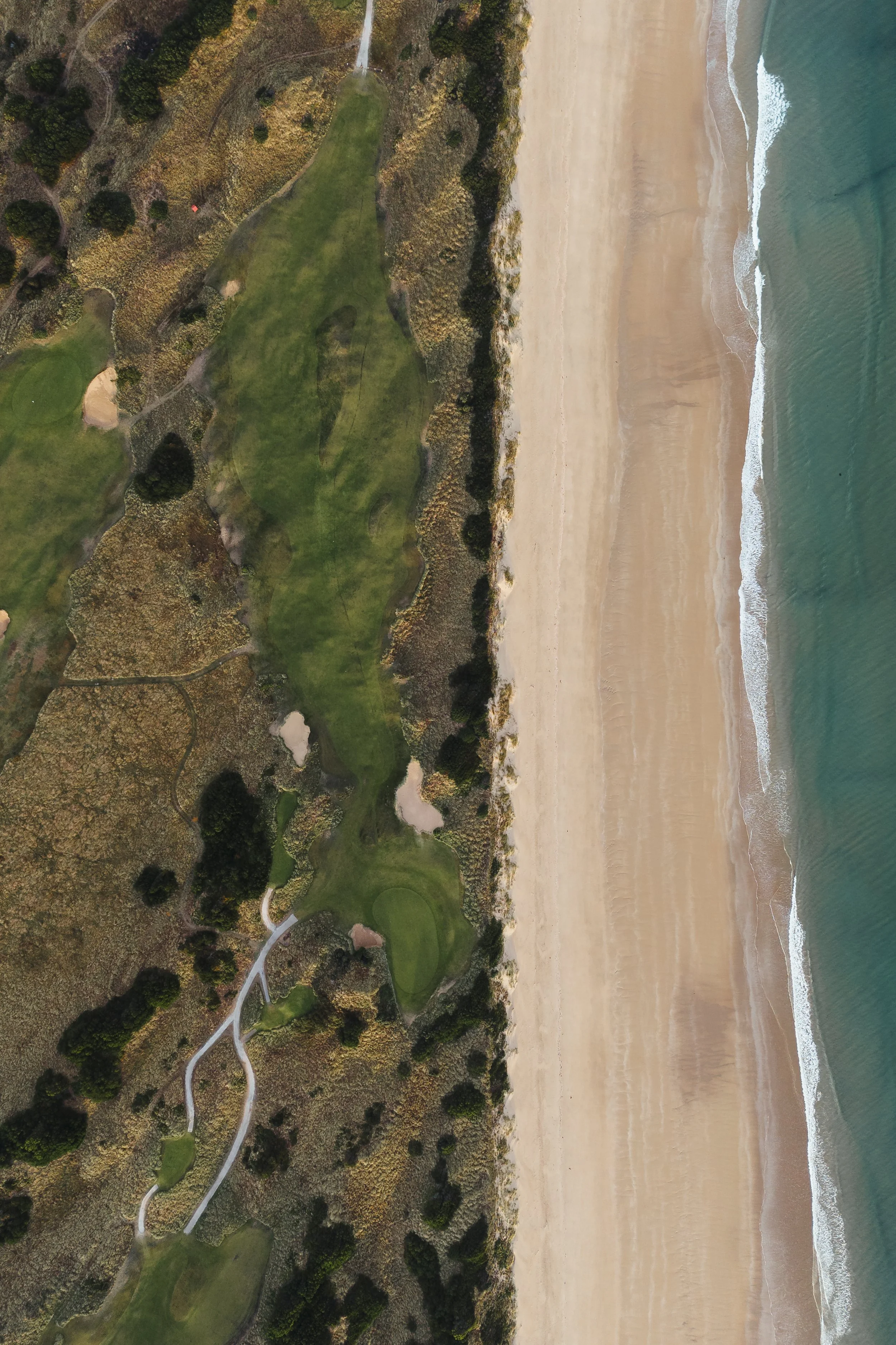 Aerial view of a coastal landscape showing a golf course with green fairways and sand bunkers running along a sandy beach with waves crashing onto the shore.