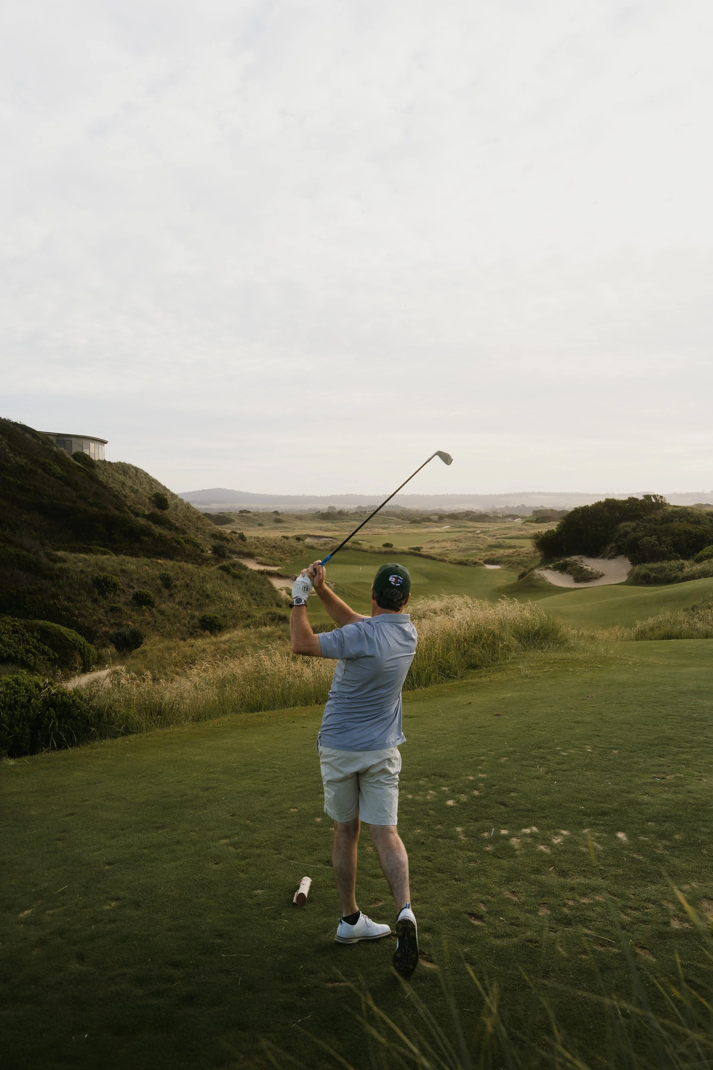 A man playing golf on a course, swinging a golf club, with hills, a pathway, and a building in the background under a cloudy sky.