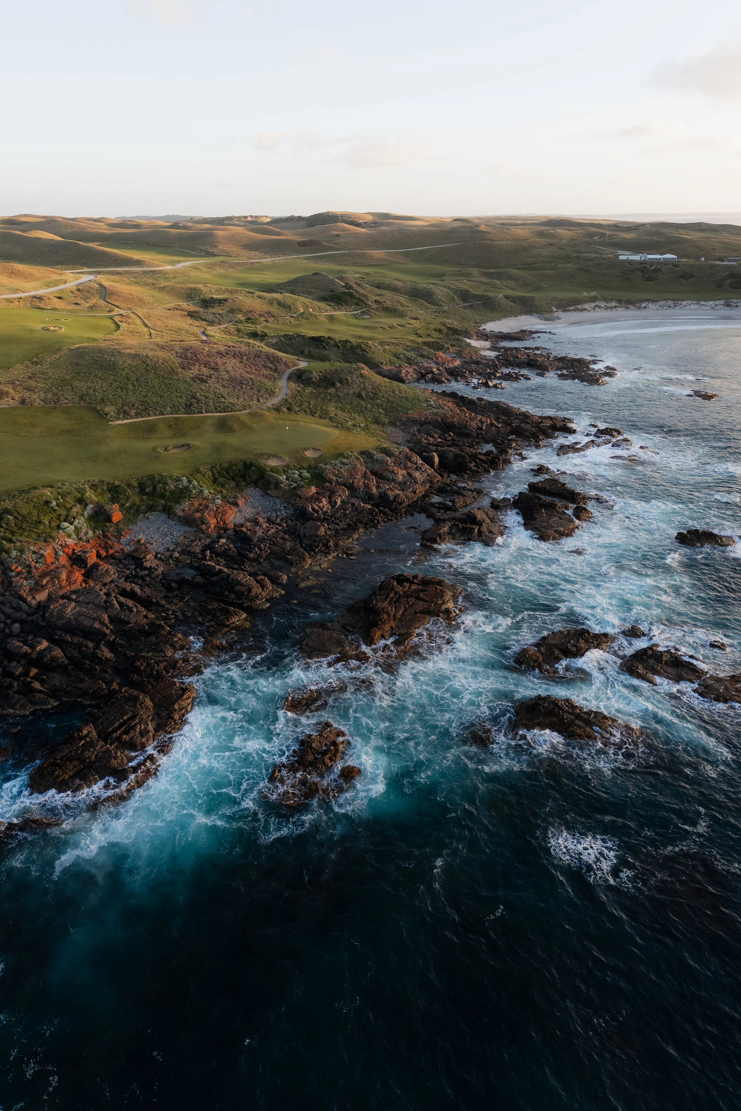 Coastal landscape featuring rocky shoreline and waves crashing against rocks, with rolling green hills and few buildings in the background under a clear sky.