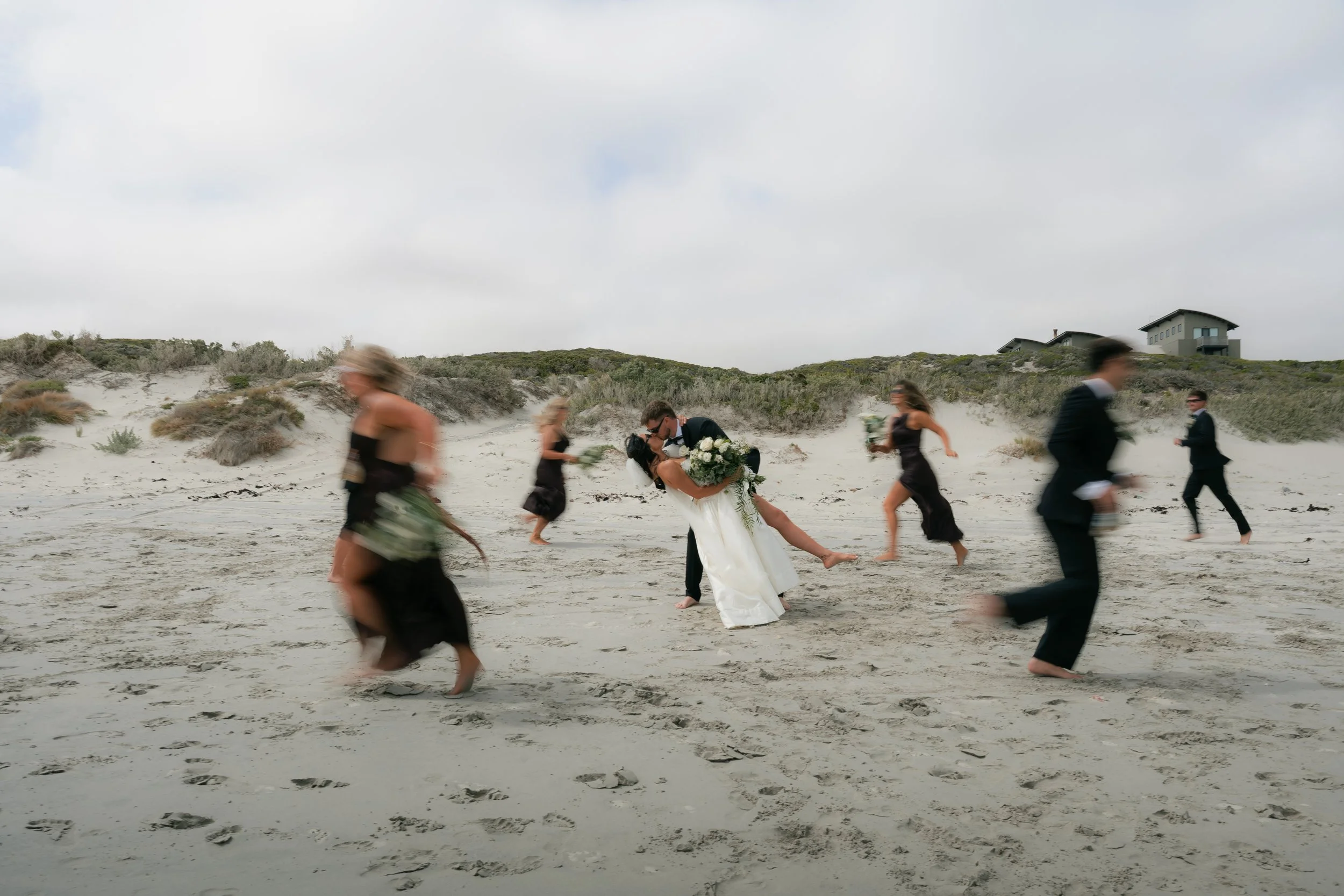 A wedding couple embrace on a sandy beach while people in formal attire walk past quickly, with houses on a hill in the background.