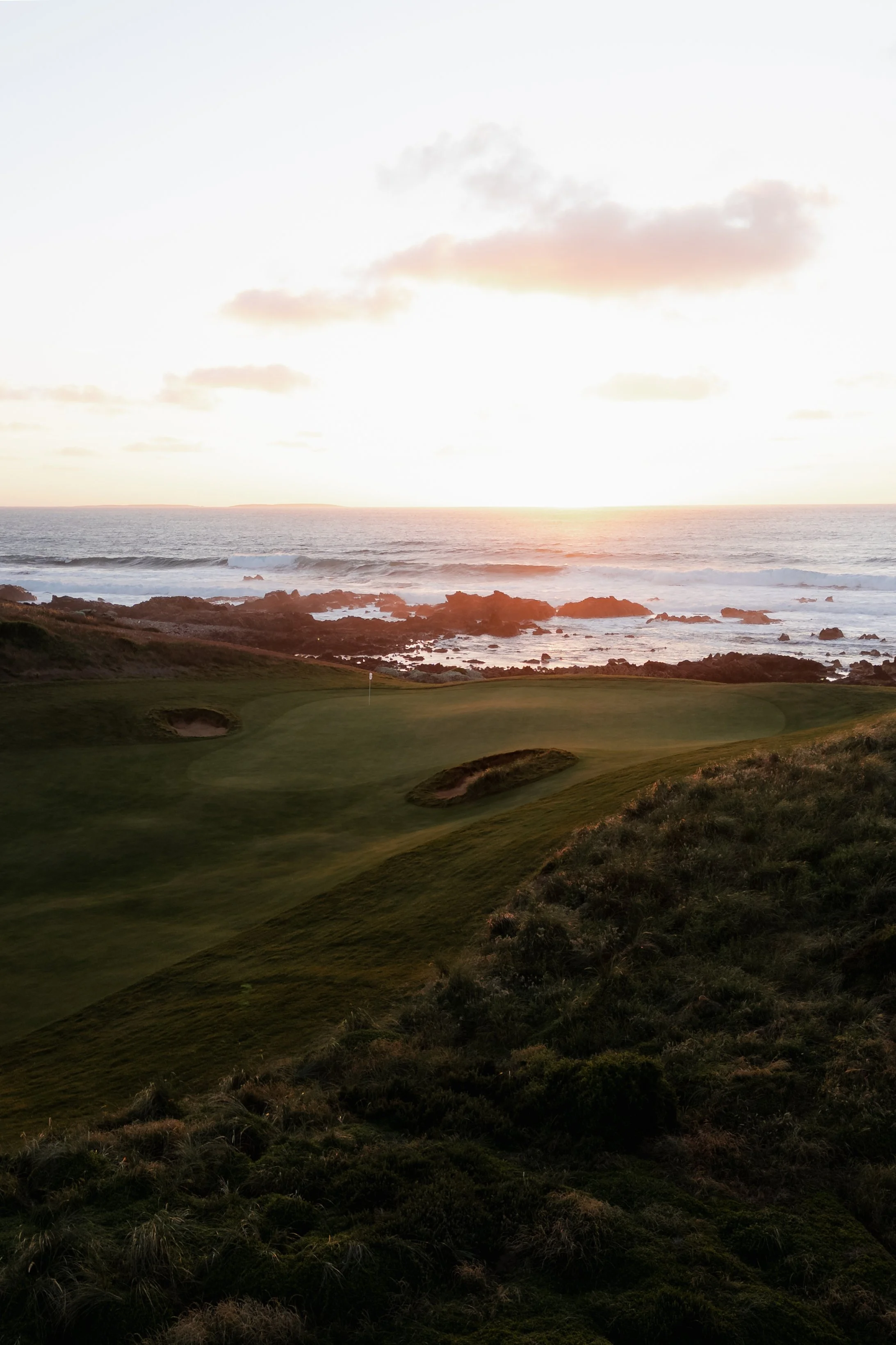 A coastal scene at sunset with a grassy golf course in the foreground, rocky shoreline, ocean waves, and a partly cloudy sky with pink and orange hues.