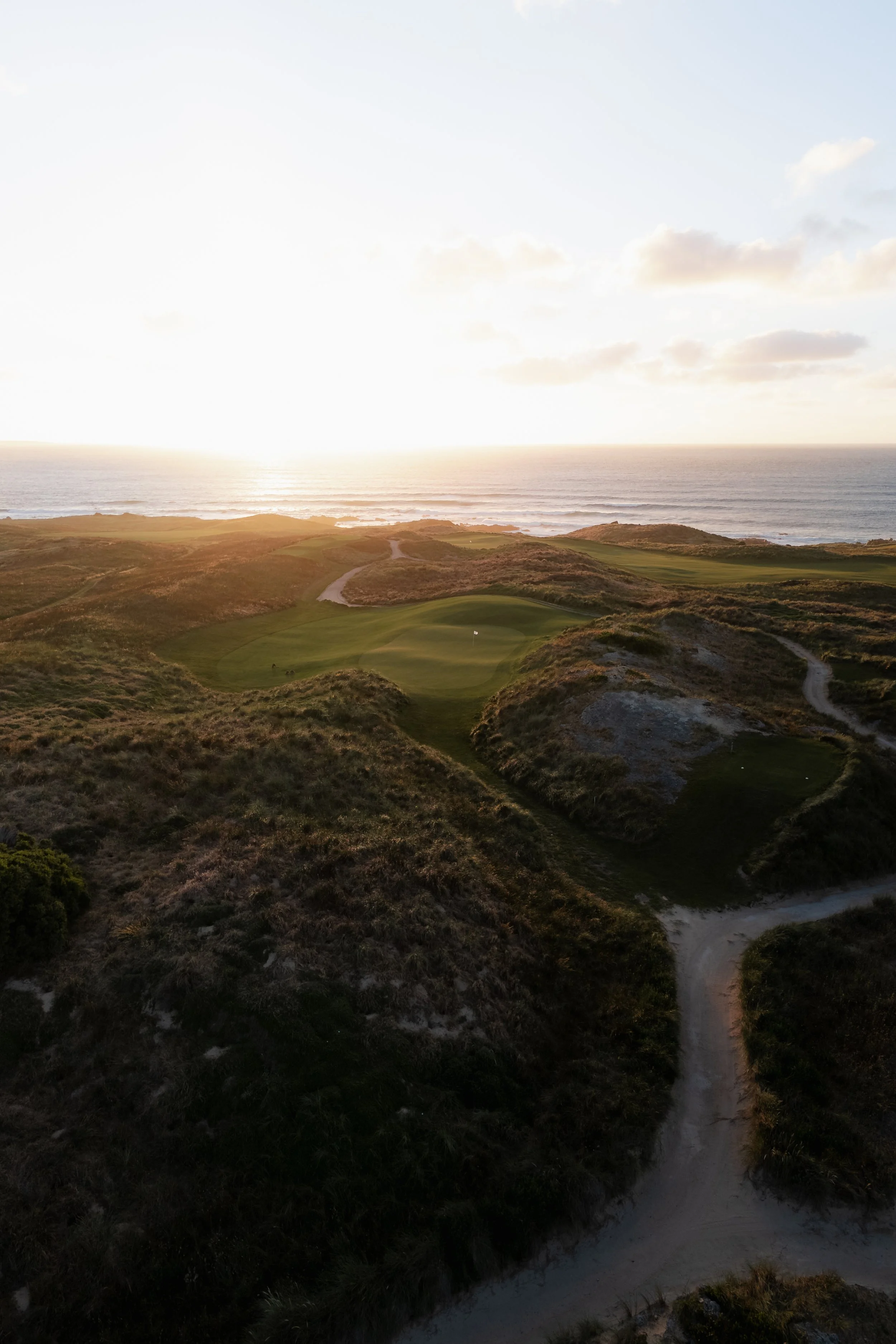 A coastal golf course with a green area, winding sand paths, and views of the ocean under a partly cloudy sky at sunset or sunrise.