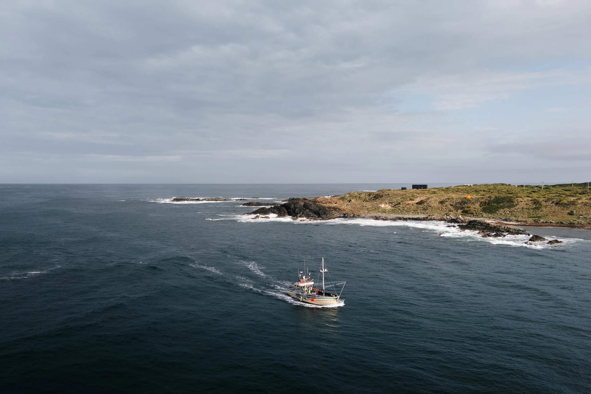 A boat sailing through the ocean near a rocky coastline with green grass and minimal structures under a cloudy sky.