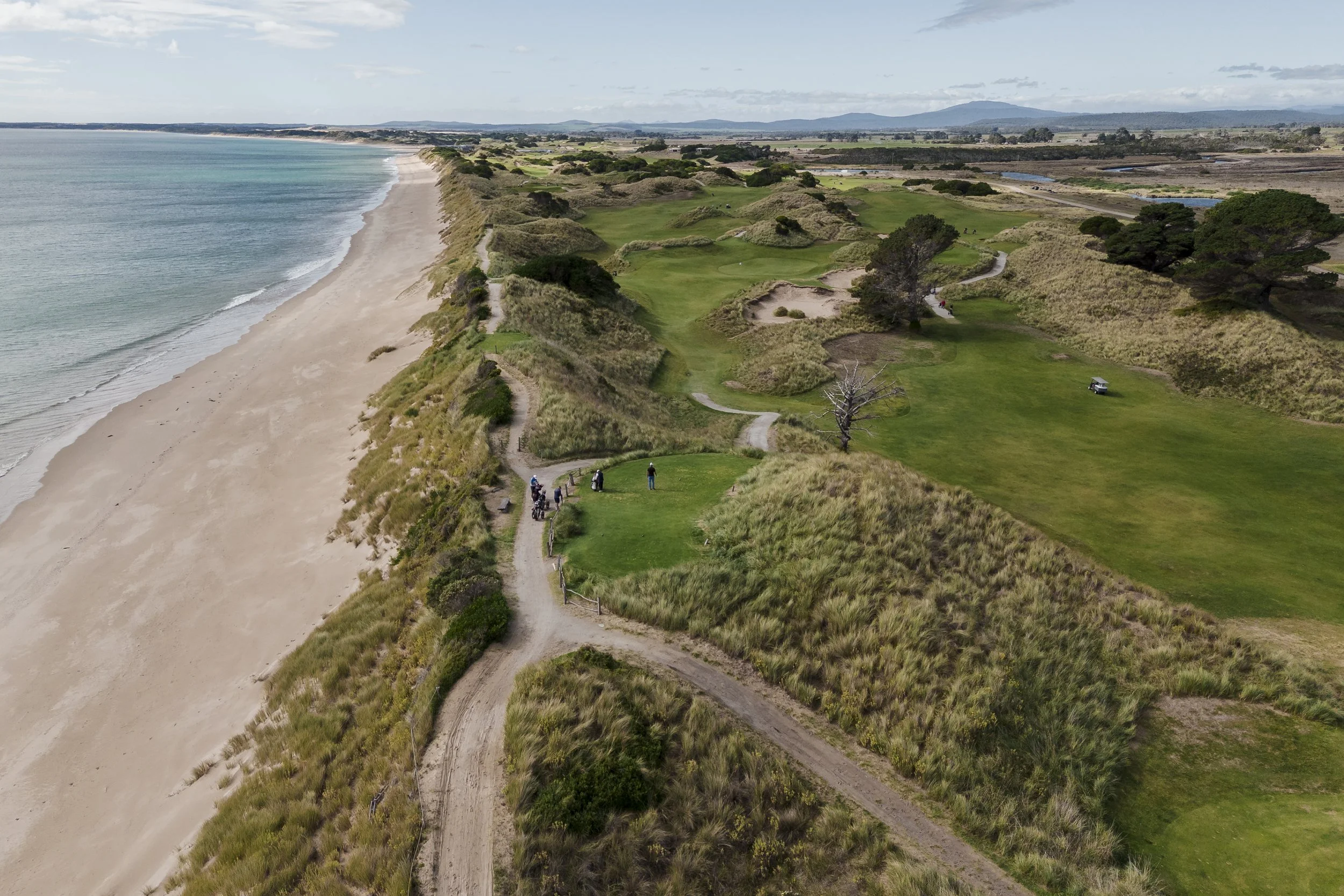 Aerial view of a coastal golf course with green fairways and sand bunkers, bordered by a sandy beach and ocean waves, with trees and distant mountains in the background.