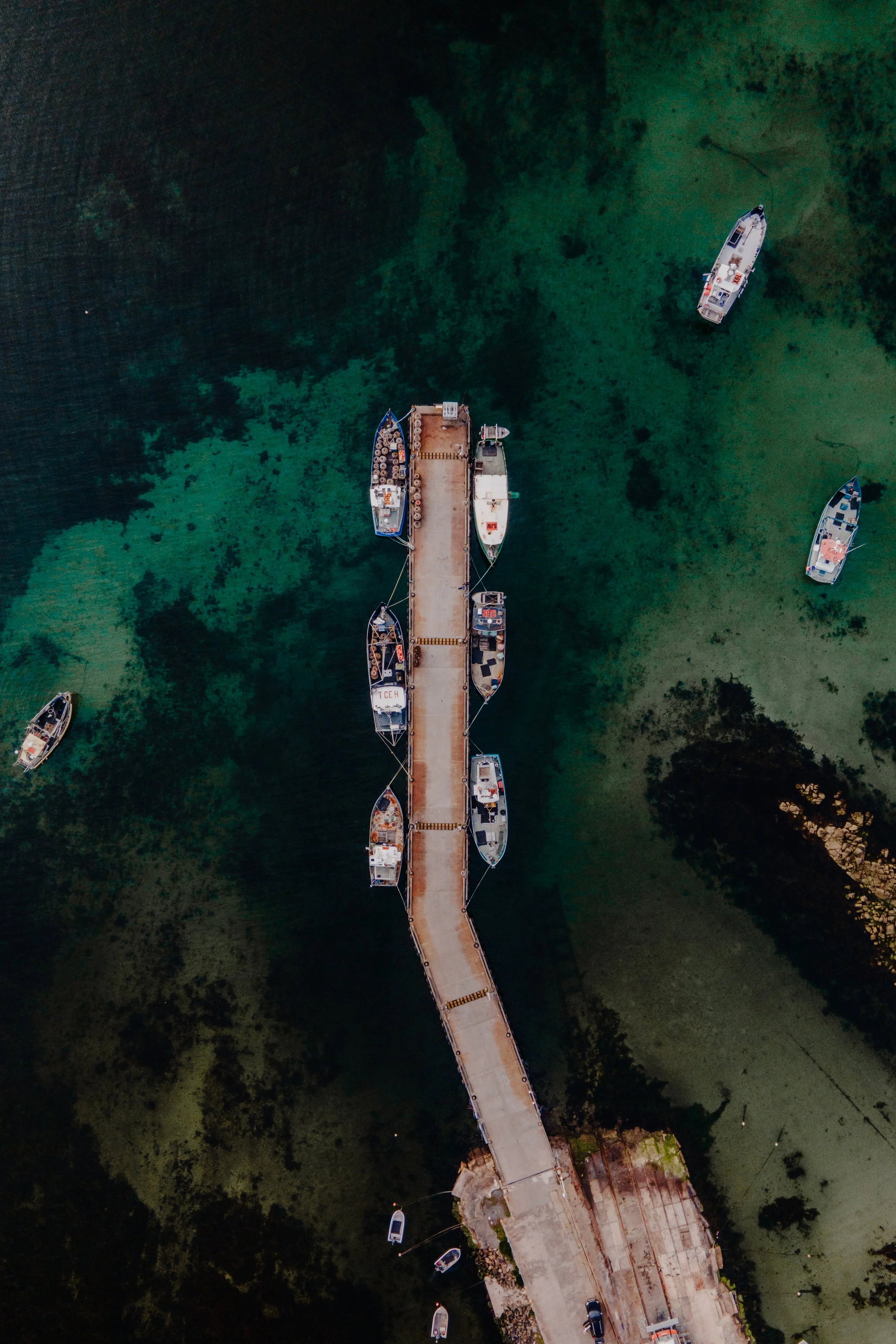 An aerial view of a narrow pier extending over green water, with several boats docked along its sides and a few small boats floating nearby.
