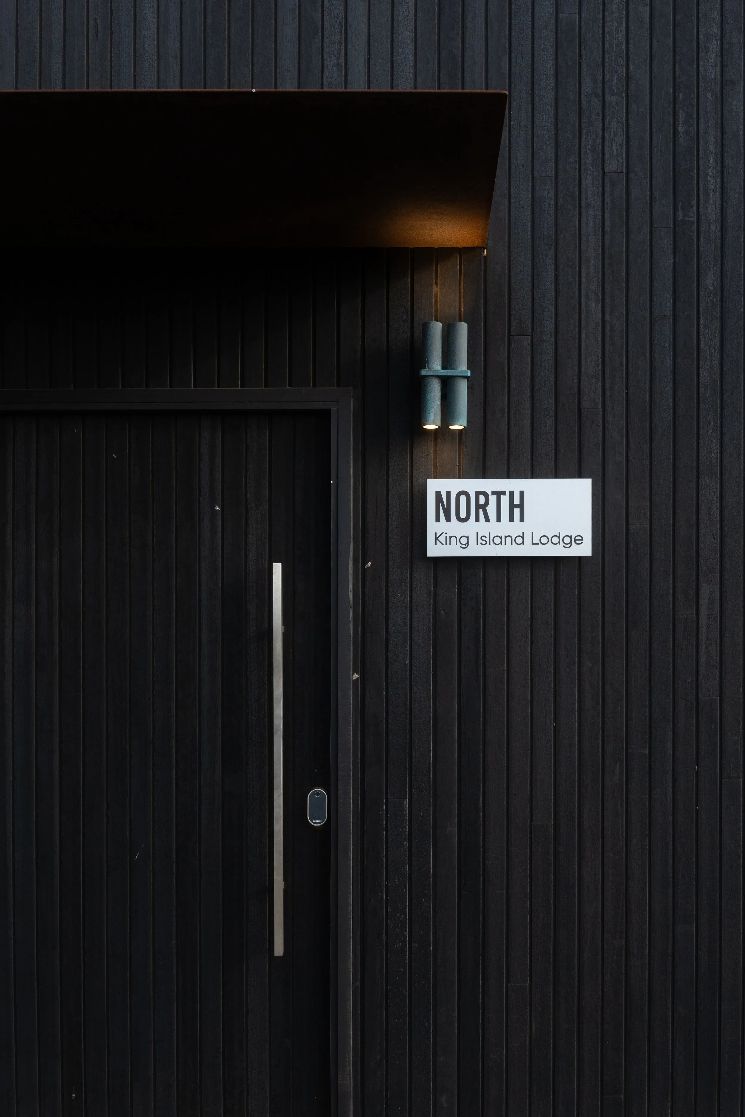 Black wooden wall with a white sign that reads 'NORTH King Island Lodge', illuminated by two small lights.