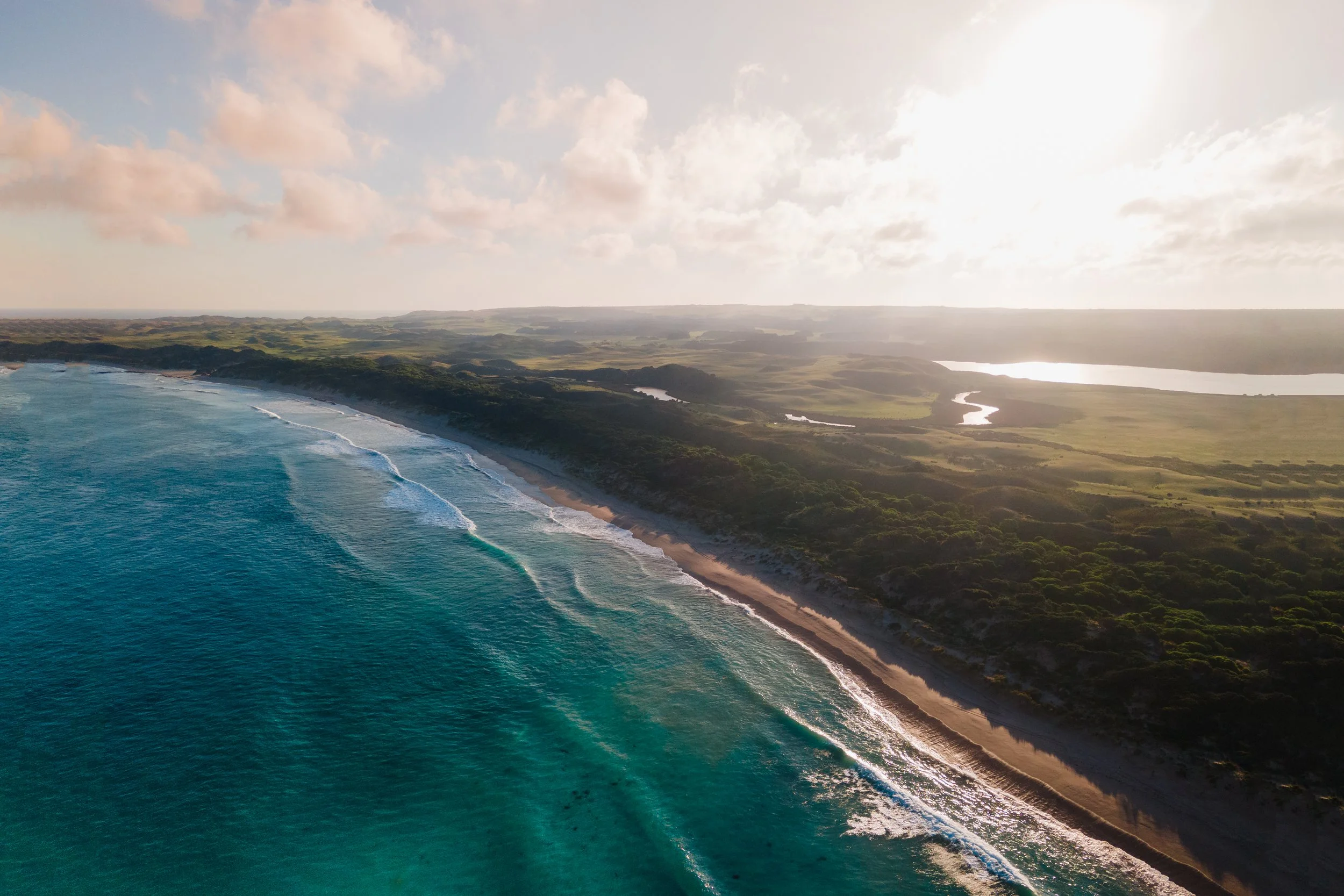 Aerial view of a coastline with blue ocean waves crashing onto a sandy beach, green forested area, and a winding river under a partly cloudy sky.
