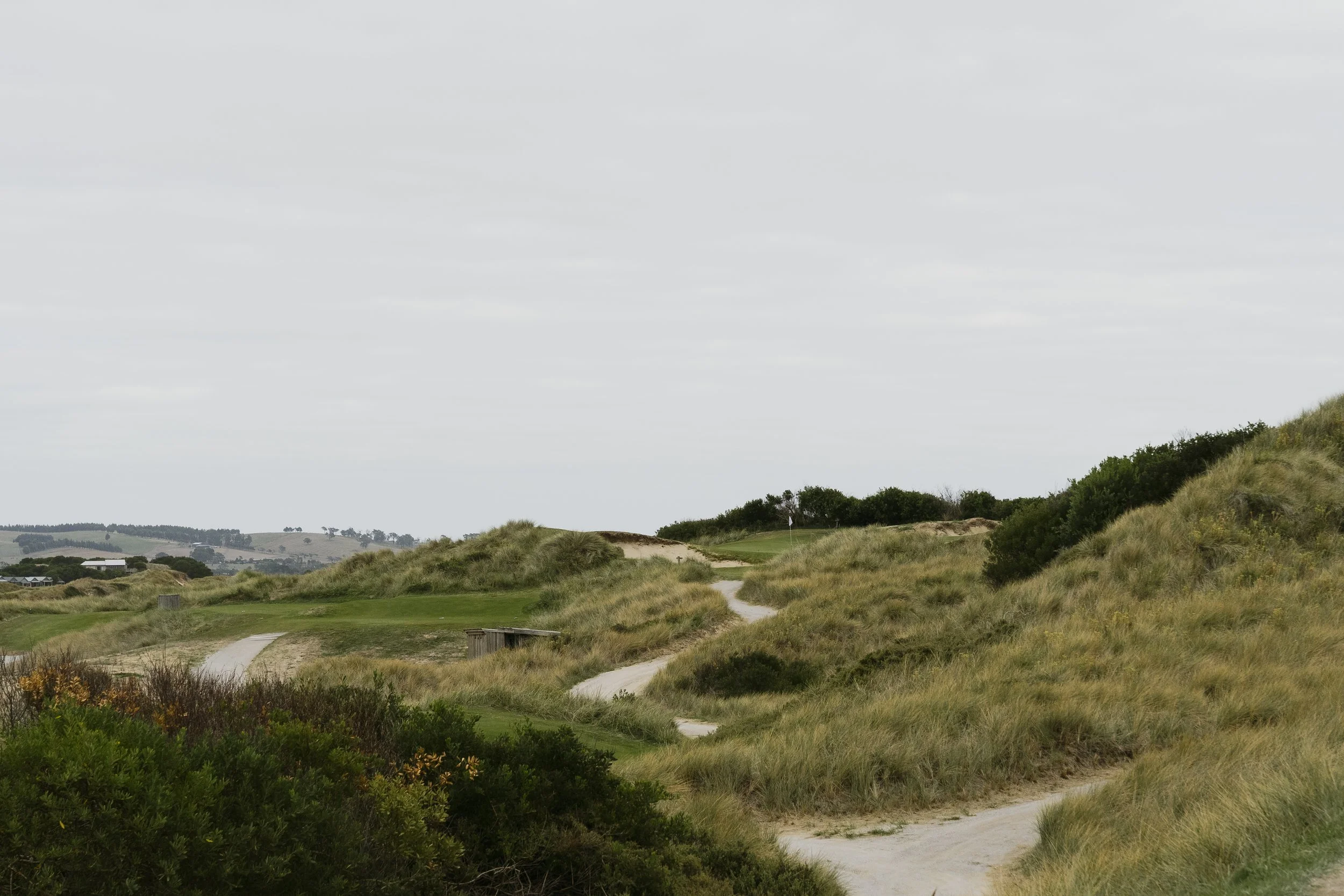 A scenic view of a golf course with a winding path surrounded by grassy hills and bushes under an overcast sky.