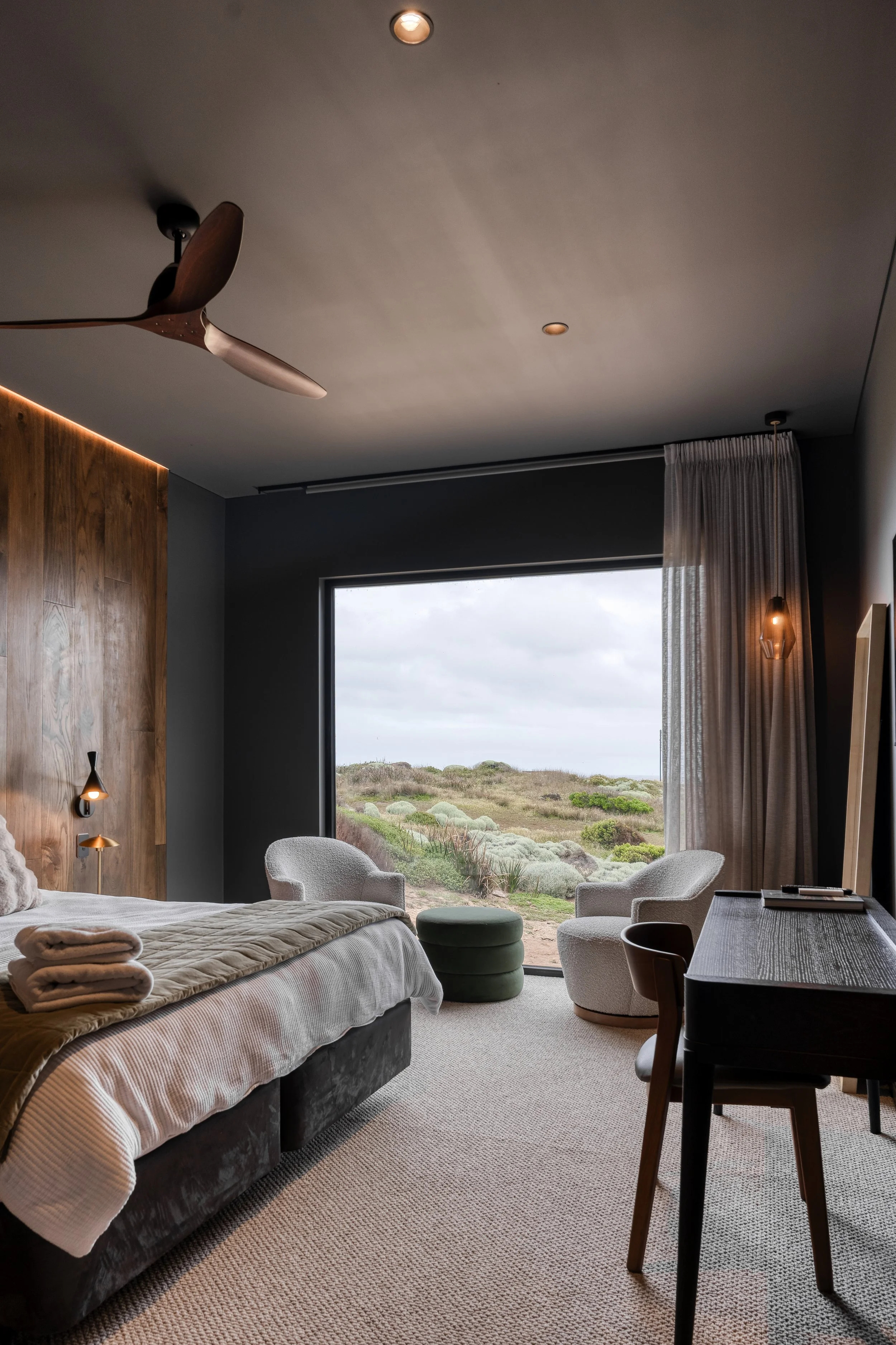 A modern bedroom with a bed, two white armchairs, a window overlooking a landscape with grass and bushes, a wooden accent wall, and ceiling lights.
