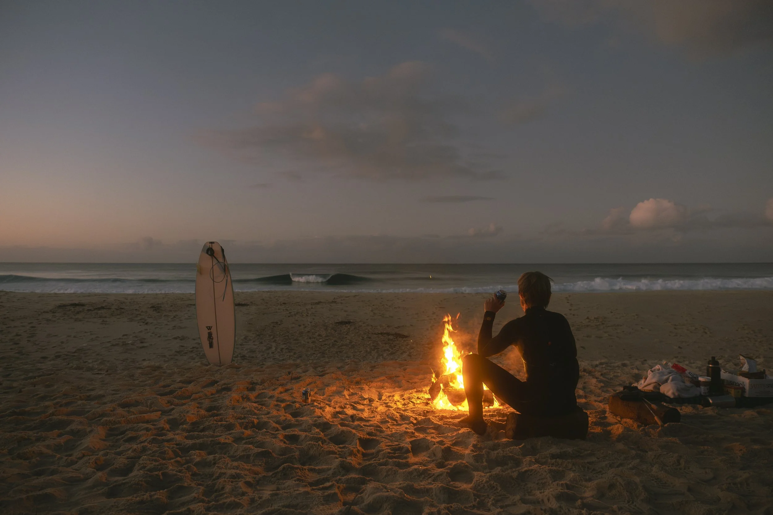 A person sitting on a sandy beach near a bonfire at sunset, holding a drink in hand, with a surfboard standing upright nearby. The ocean waves are visible in the background, and the sky is partly cloudy.