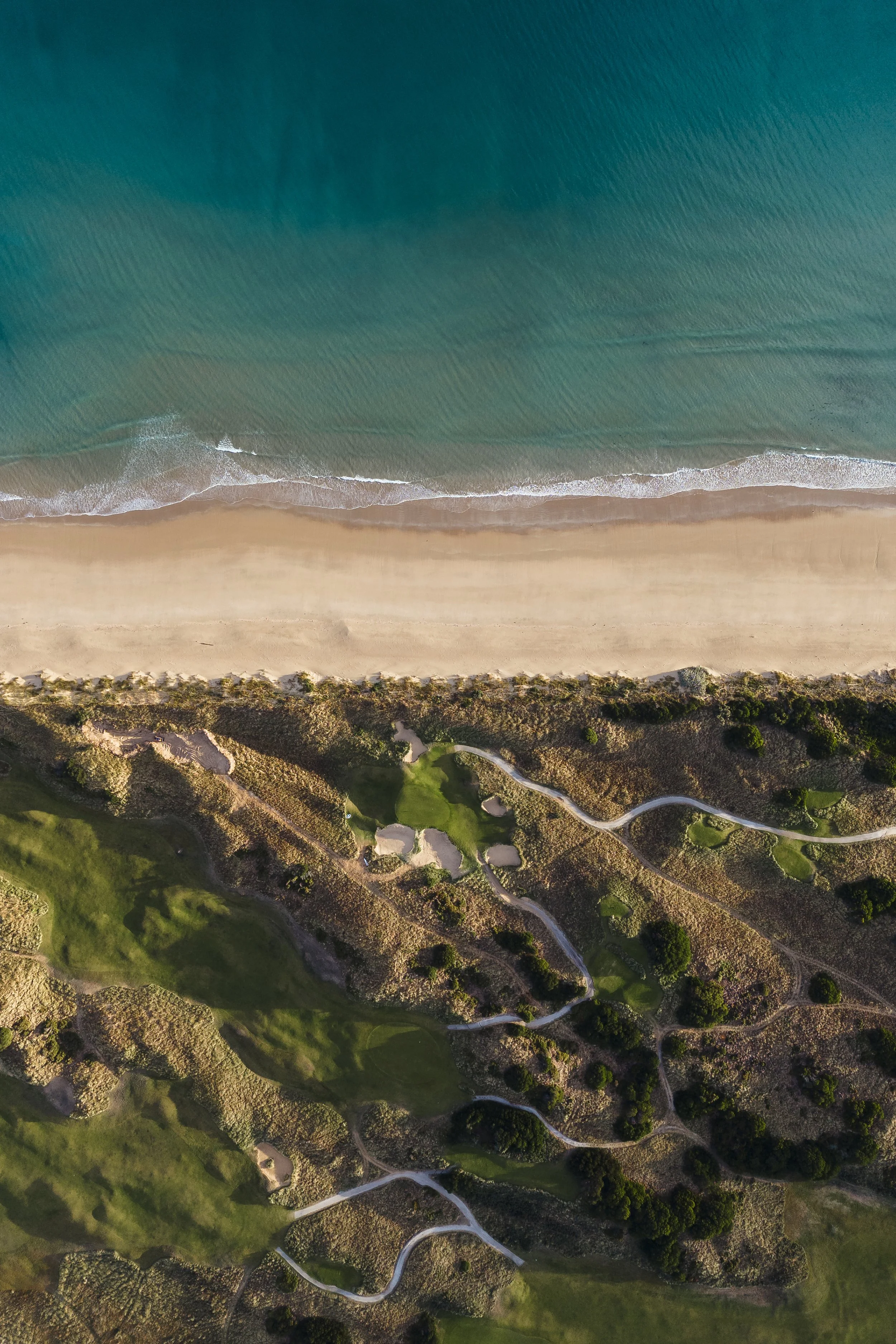 An aerial view of a coastline with turquoise ocean waves gently crashing onto a sandy beach. The beach is bordered by greenery and a winding pathway.