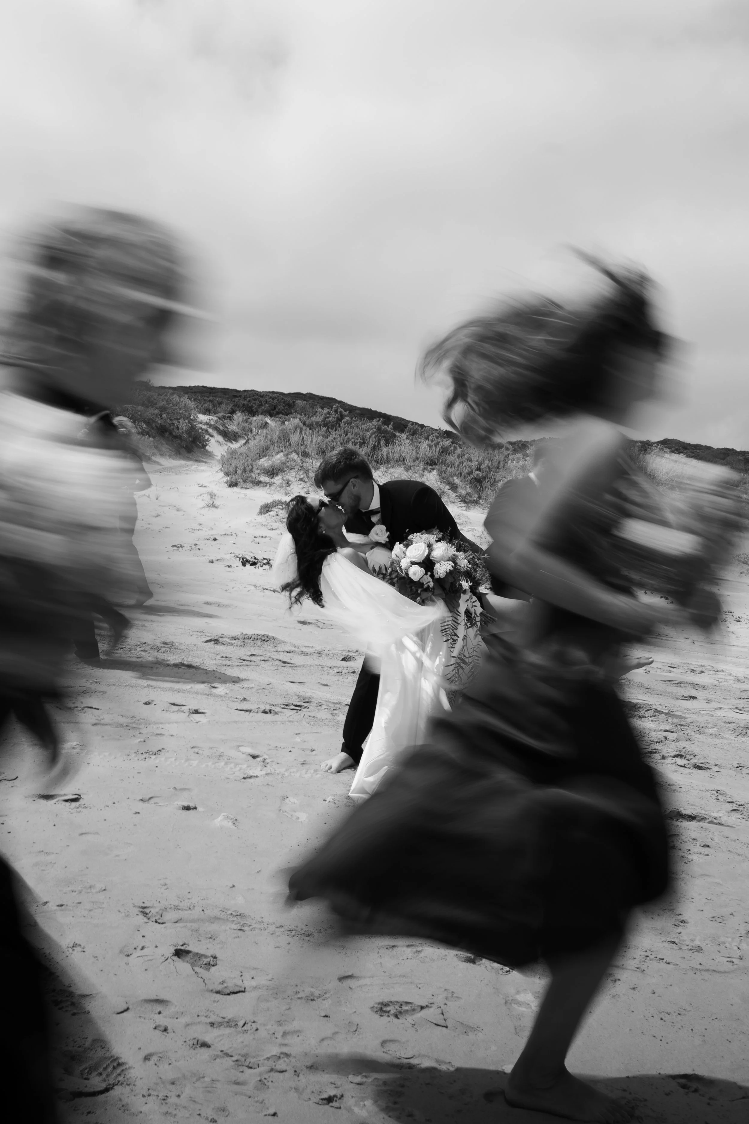 A couple dressed in wedding attire sharing a kiss on a sandy beach, surrounded by blurred figures of people walking, with dunes and cloudy sky in the background.
