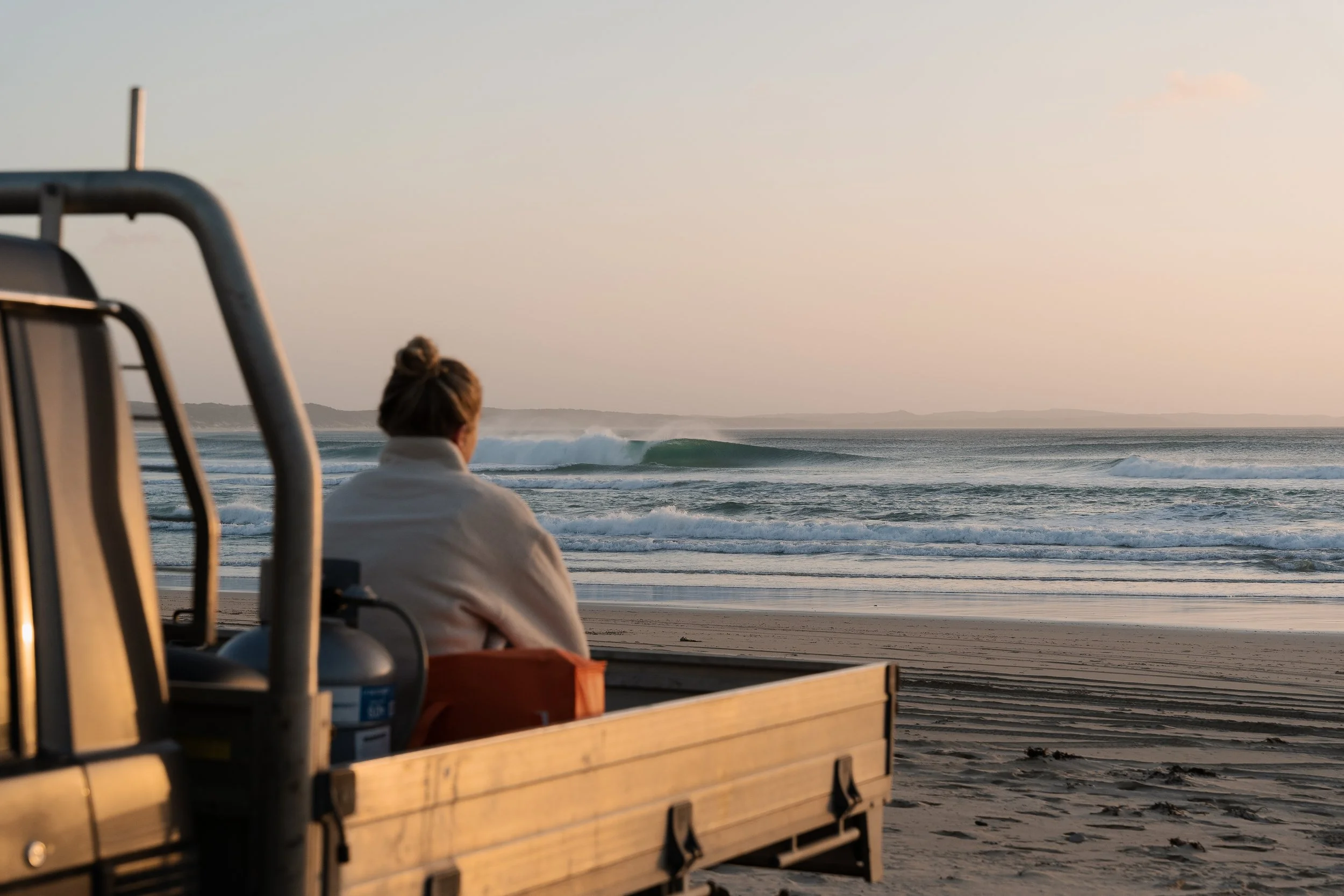 Person sitting on a bench at the beach, facing the ocean during sunset with gentle waves and distant land in the background.