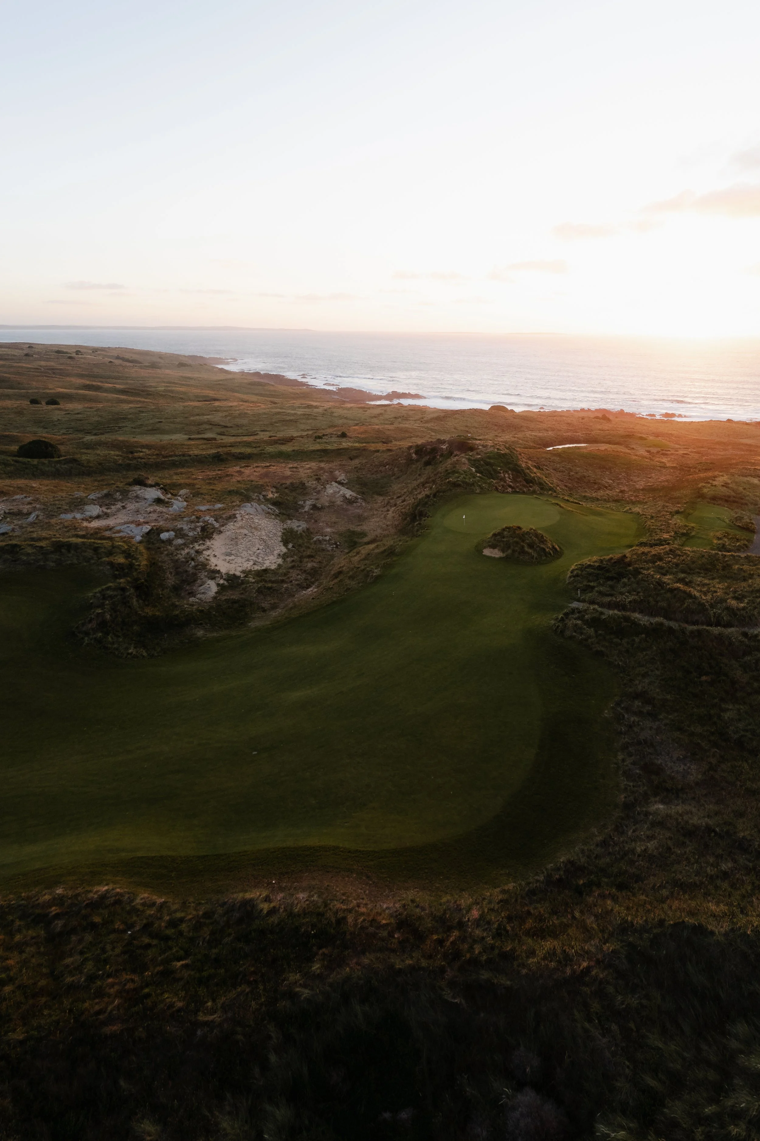 A scenic view of a golf course along a coastline at sunset, with green fairways, rugged terrain, and the ocean in the background.