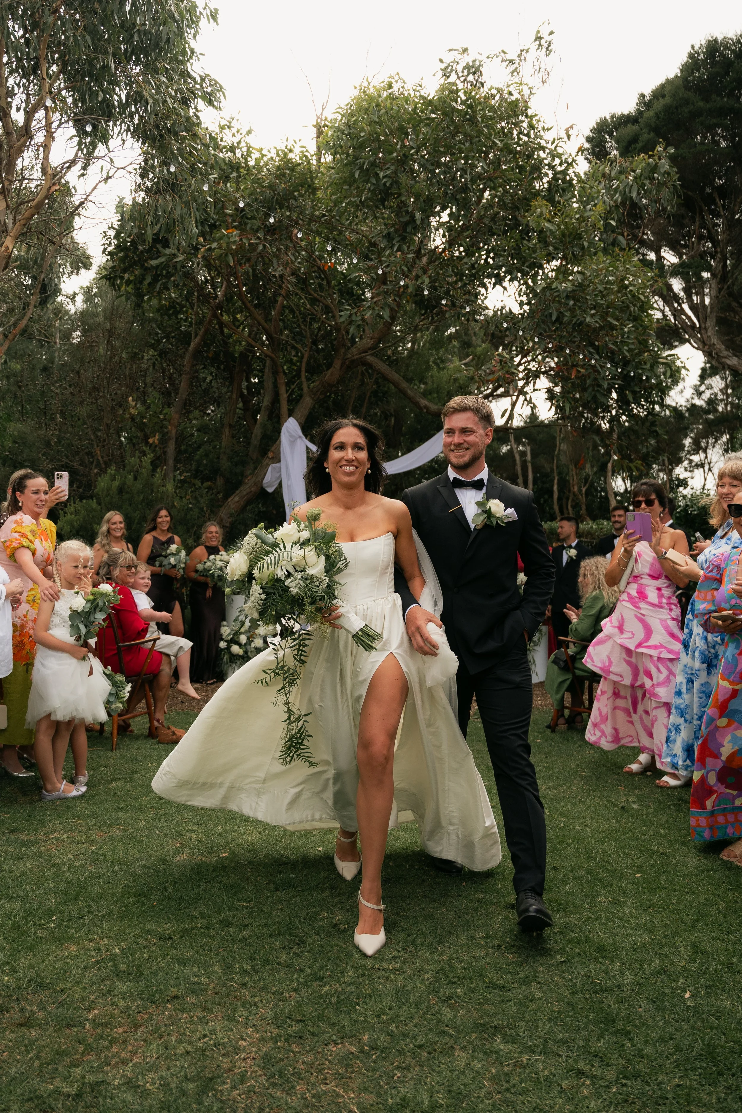 A bride and groom walking down the aisle at an outdoor wedding ceremony, surrounded by guests and decorated with string lights and trees.