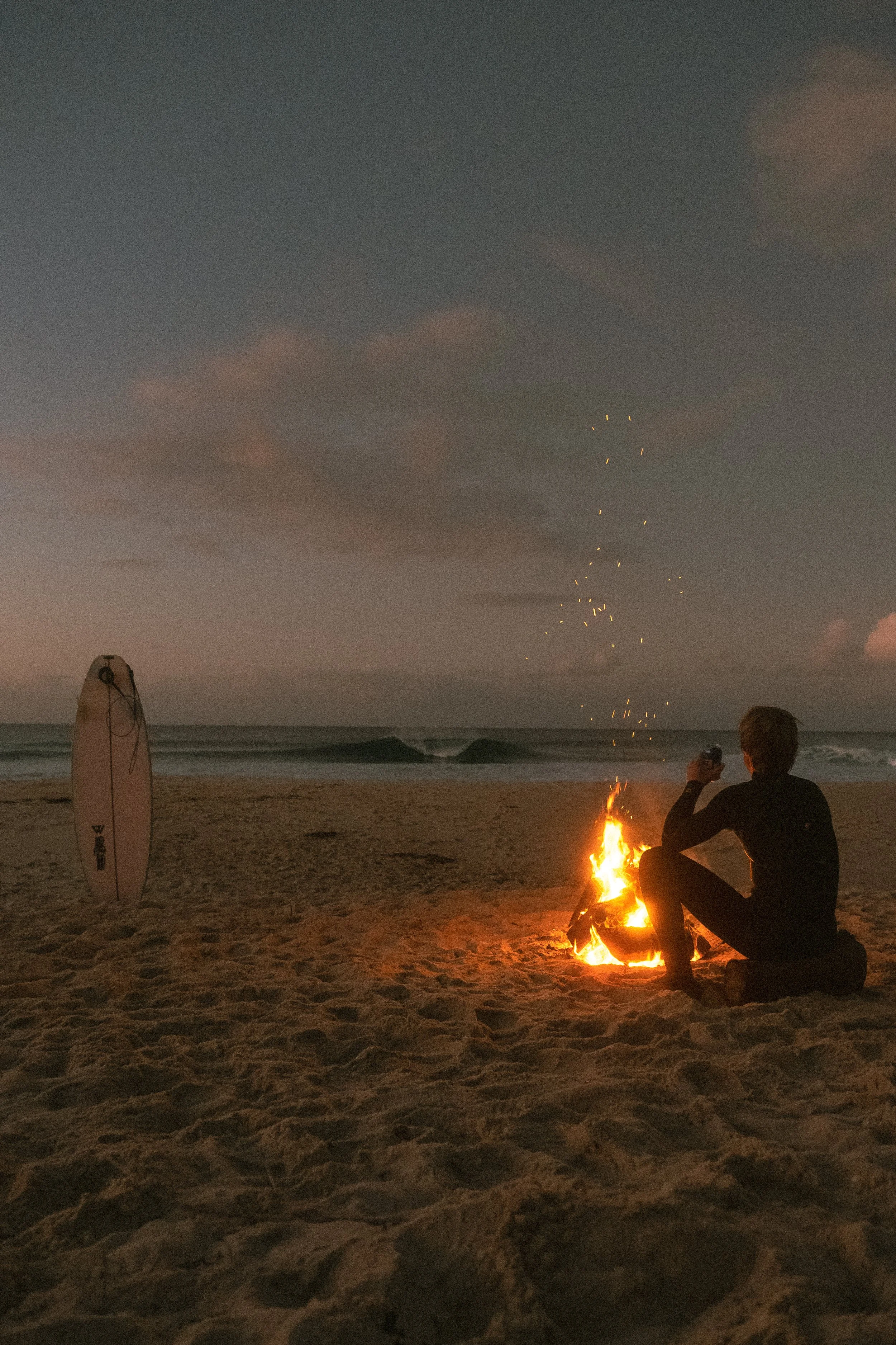 Person sitting on the sand by a small campfire on the beach during dusk, with a surfboard standing upright nearby and a cloudy sky with faint sparkles or stars in the background.