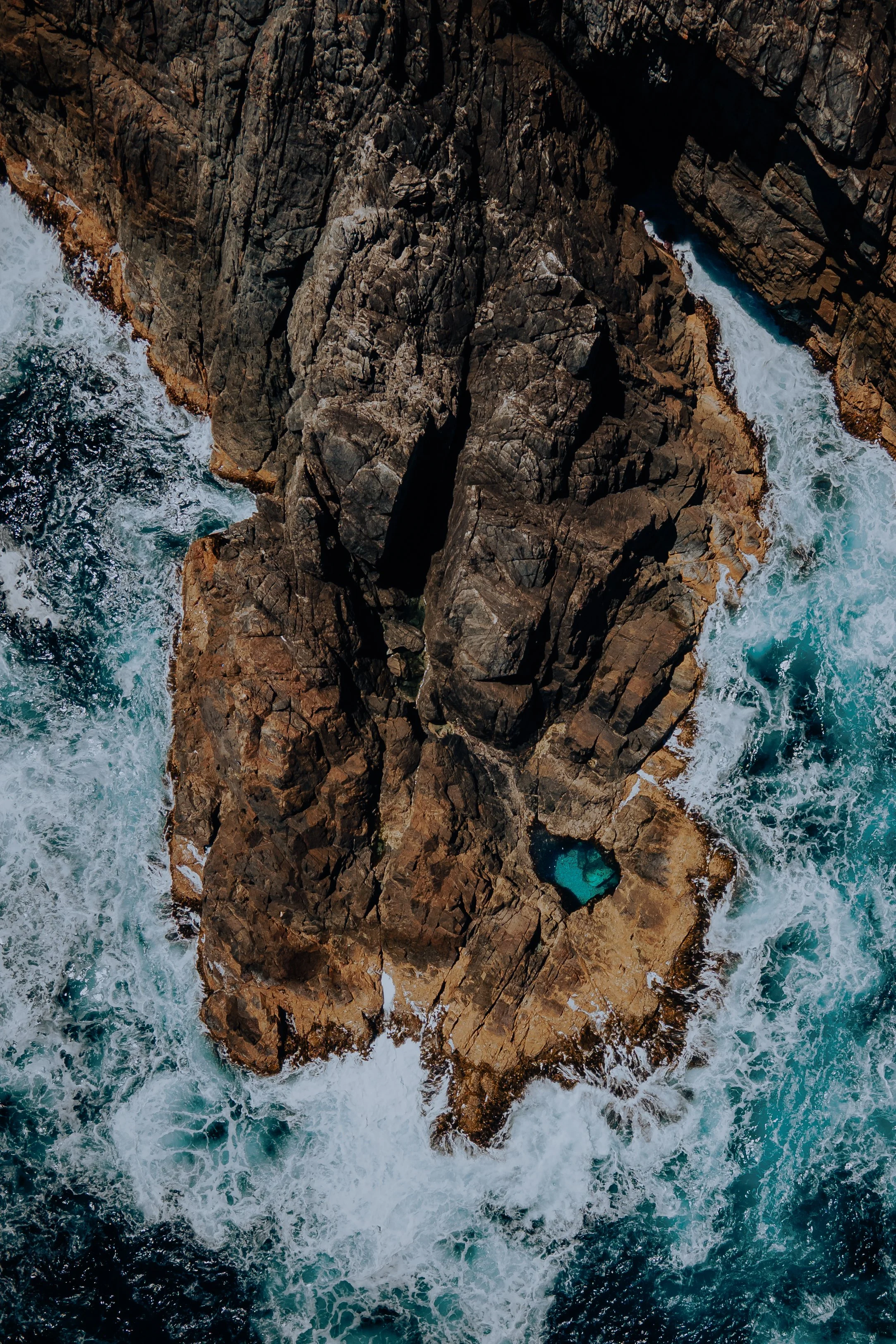 An aerial view of rugged rocky cliffs in the ocean, with waves crashing against the rocks and a small pool of water on the rocks.