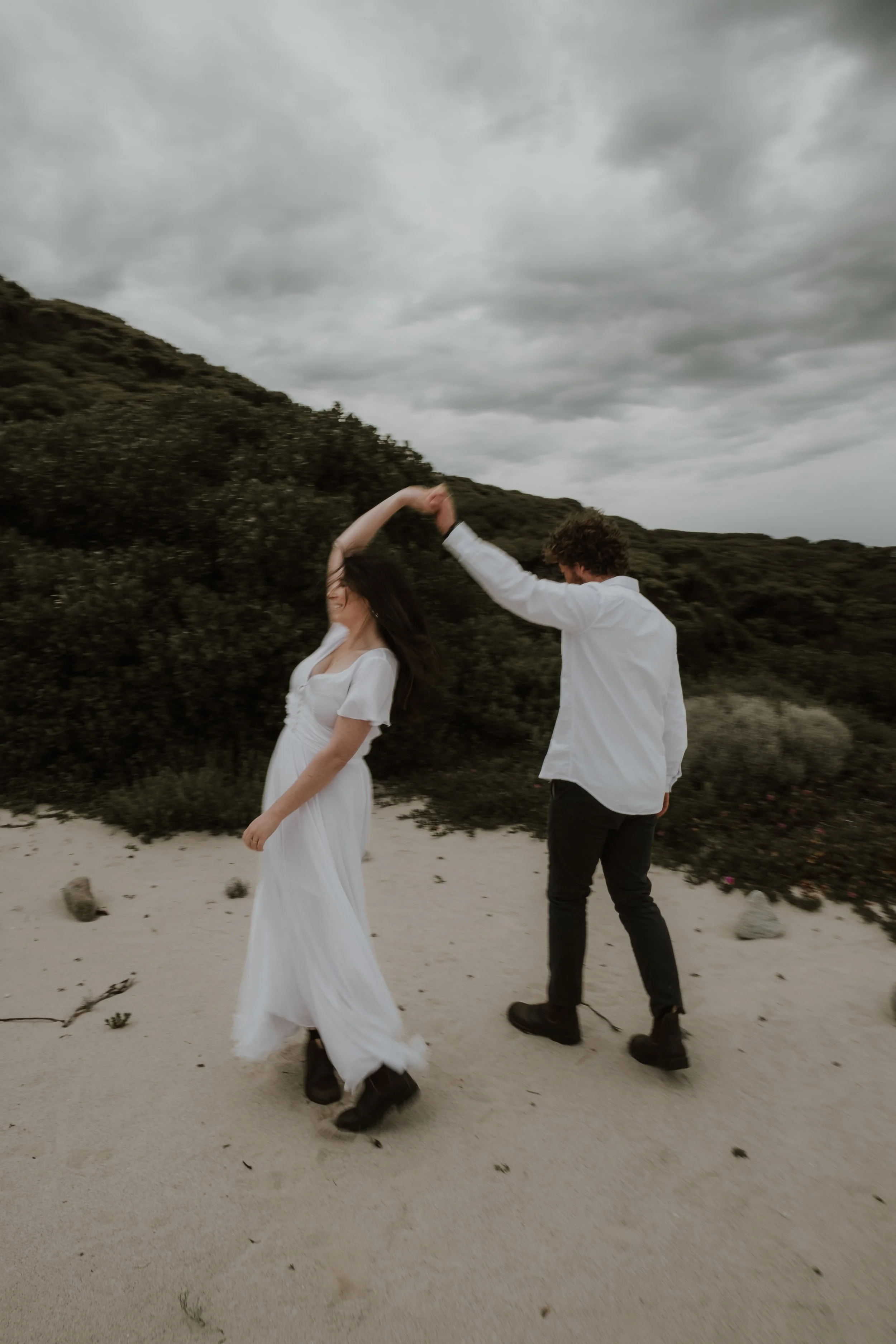 A couple dancing on a sandy beach with cloudy skies and green hills in the background.