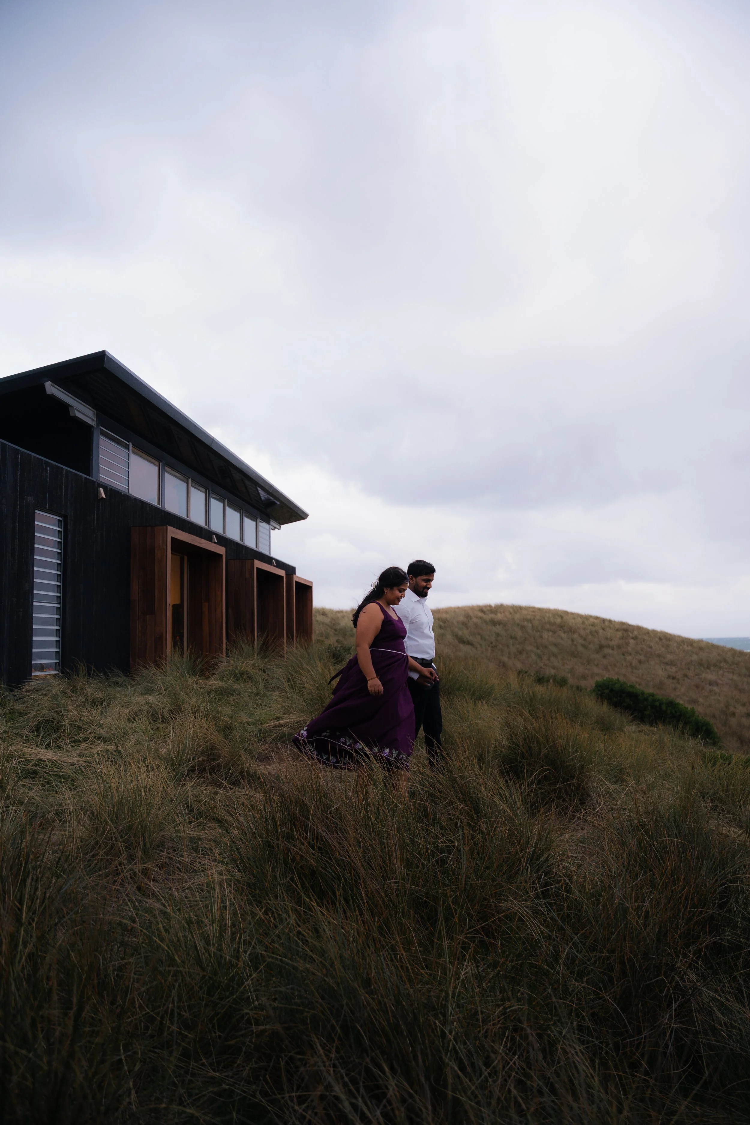 A man and woman dressed in formal attire walking through tall grass outside a modern house on a cloudy day.