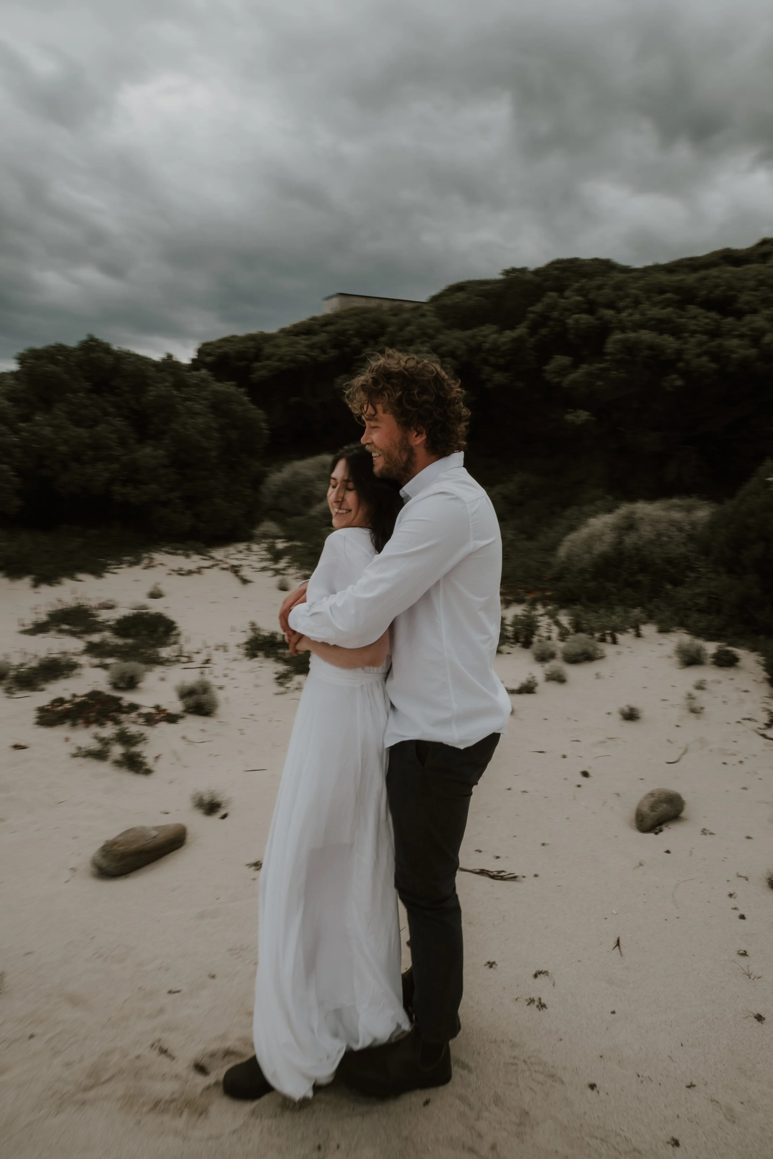 A couple embracing on a sandy beach during cloudy weather, with green bushes and trees in the background.