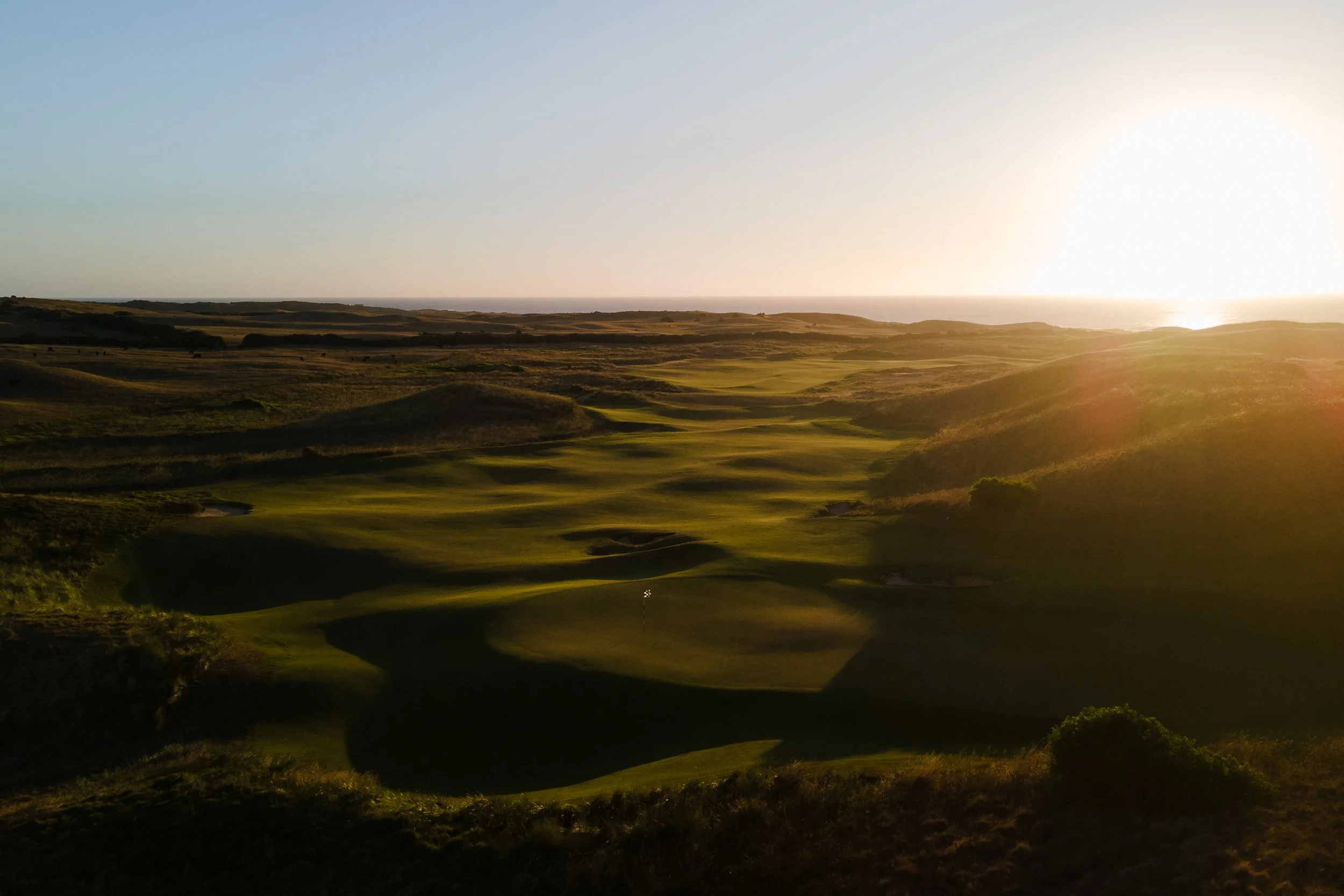 A scenic view of a golf course with green fairways and sand bunkers, set against a landscape with rolling hills and an ocean in the distance, during sunset.