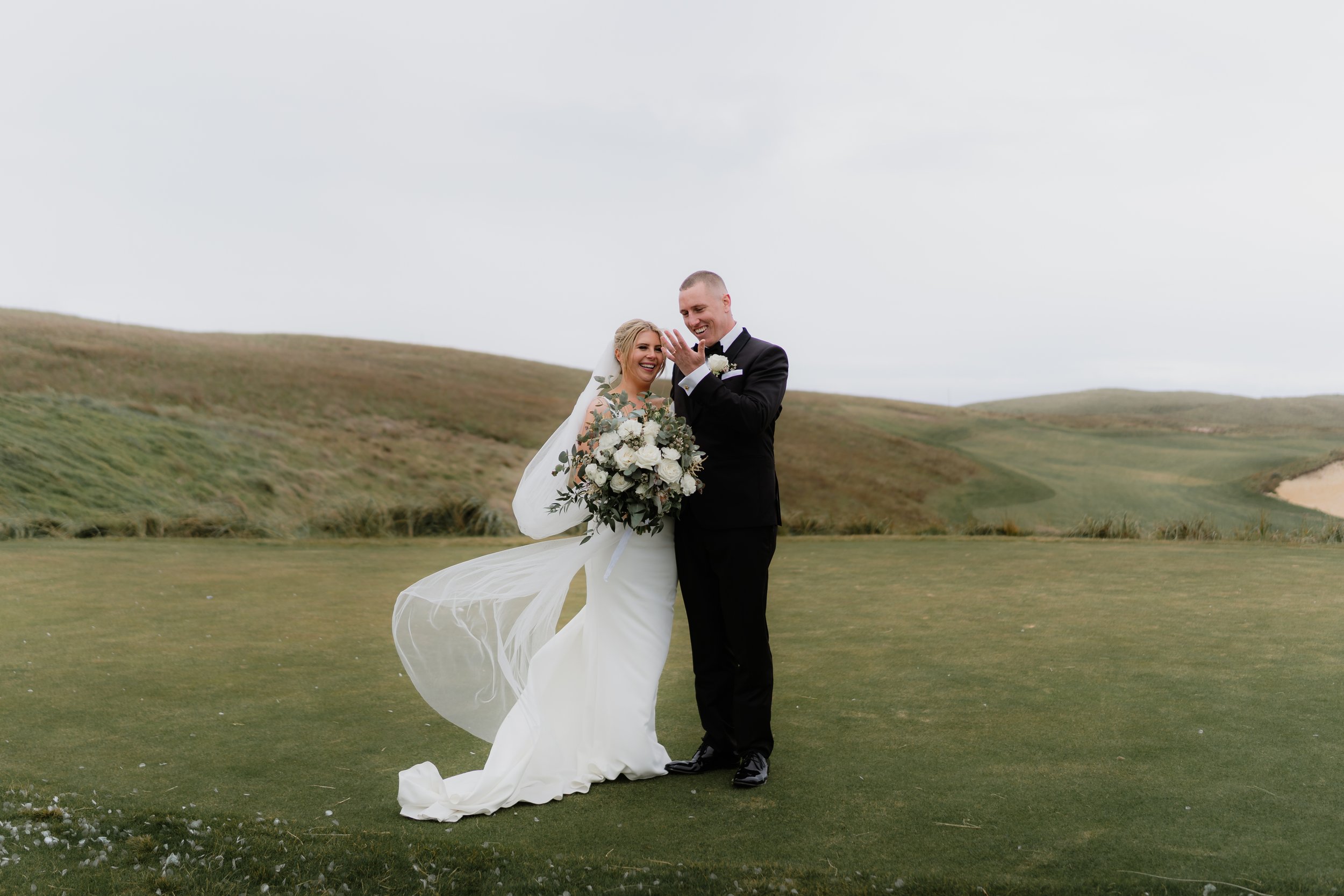 A bride and groom smiling and embracing on a grassy field with rolling hills in the background. The bride is holding a large bouquet of white and green flowers, wearing a white wedding gown and veil. The groom is dressed in a black tuxedo with a whit