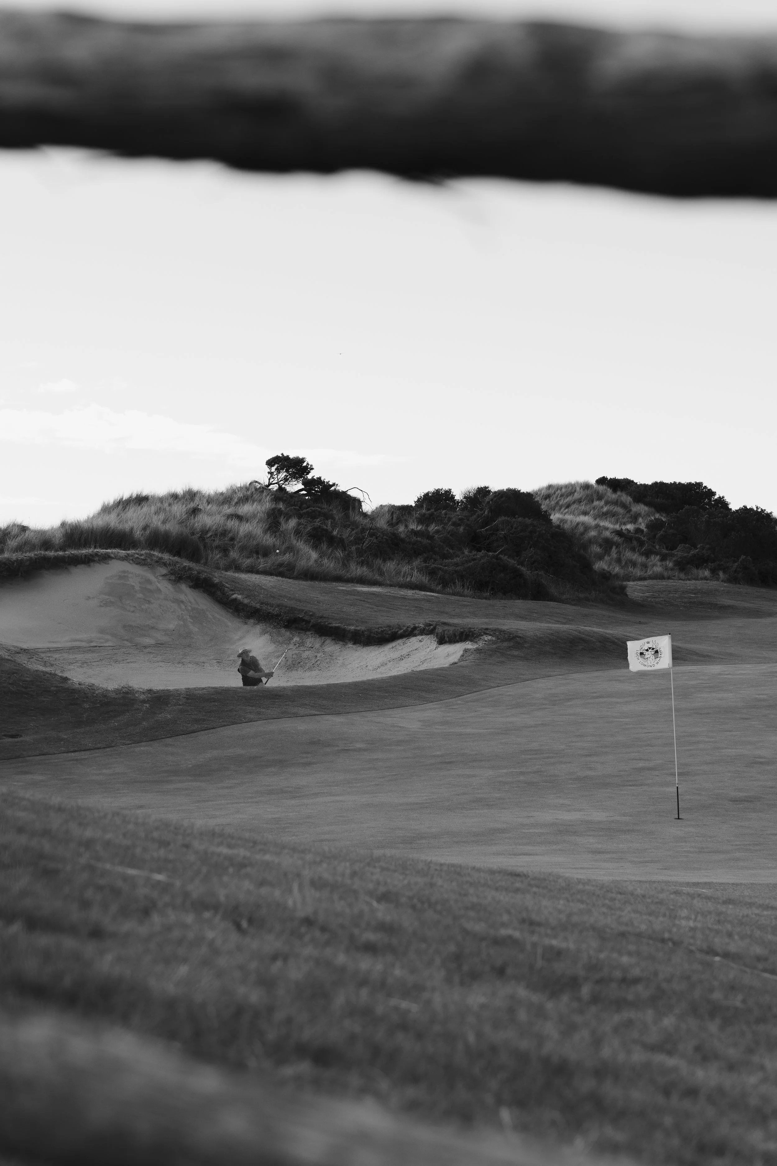 A golfer is taking a shot from a sand trap on a golf course with a flagstick and hole in the foreground. There are trees and a grassy hillside in the background.