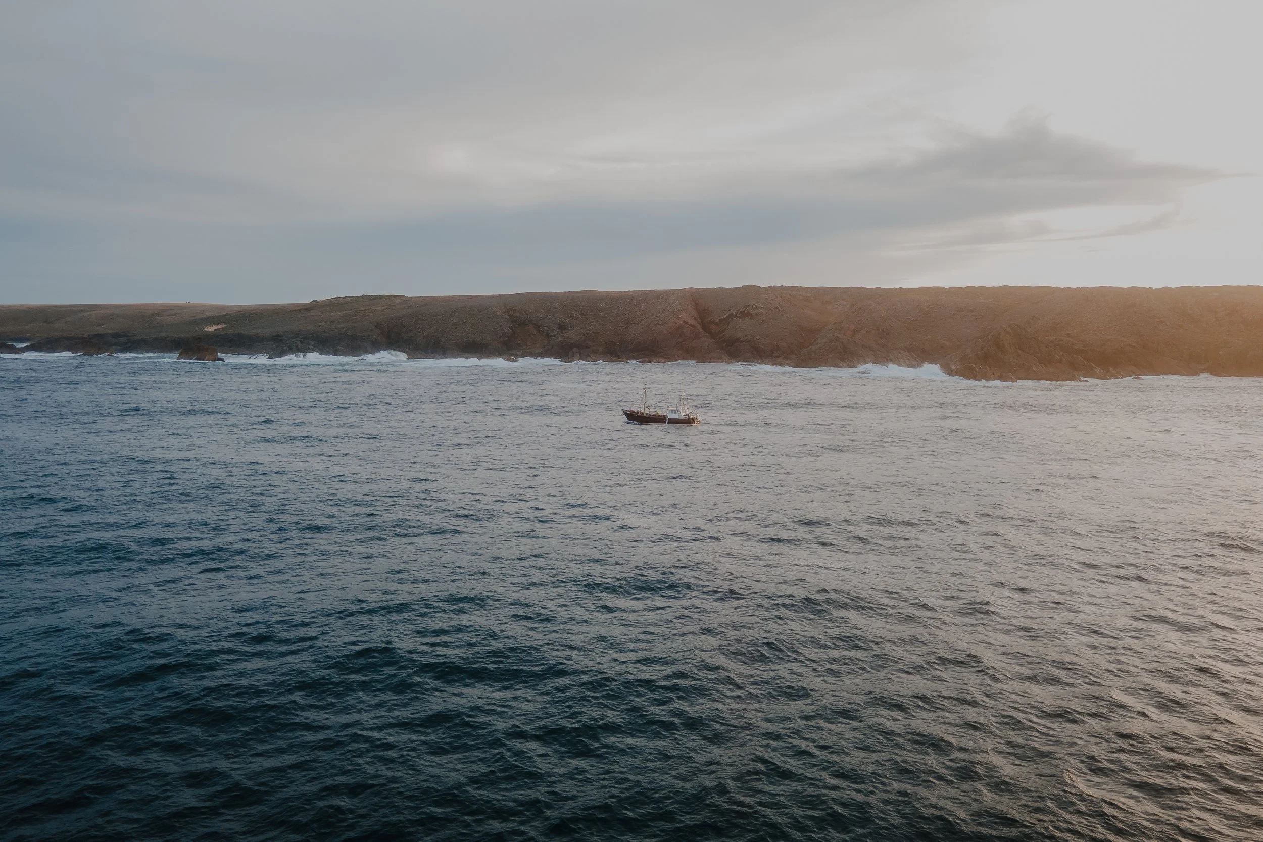 A boat floating on the ocean near rocky cliffs under a cloudy sky.