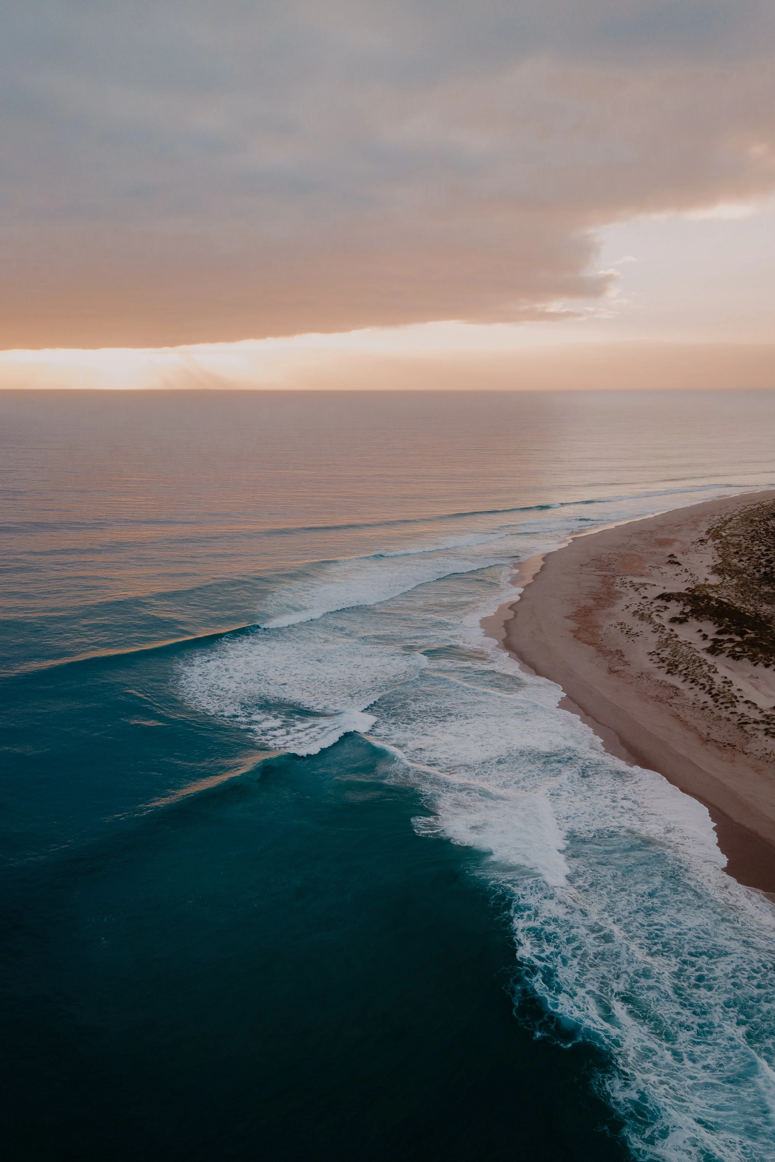 Sunset over the ocean with waves crashing onto a sandy beach and a cloudy sky in the background.