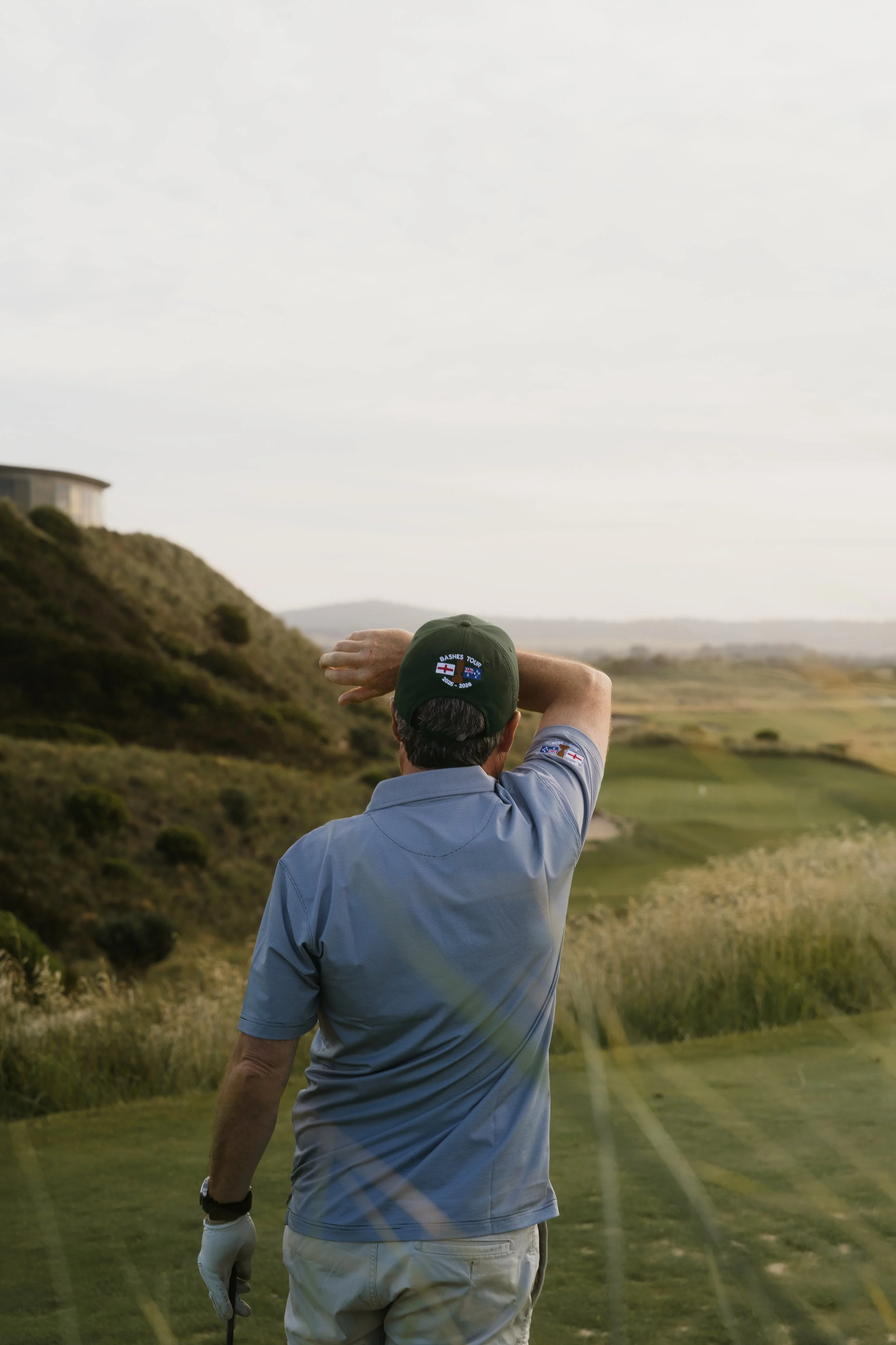 A man on a golf course, wearing a cap and a blue shirt, is holding a golf club and walking away from the camera. The landscape features rolling hills and some buildings in the distance.