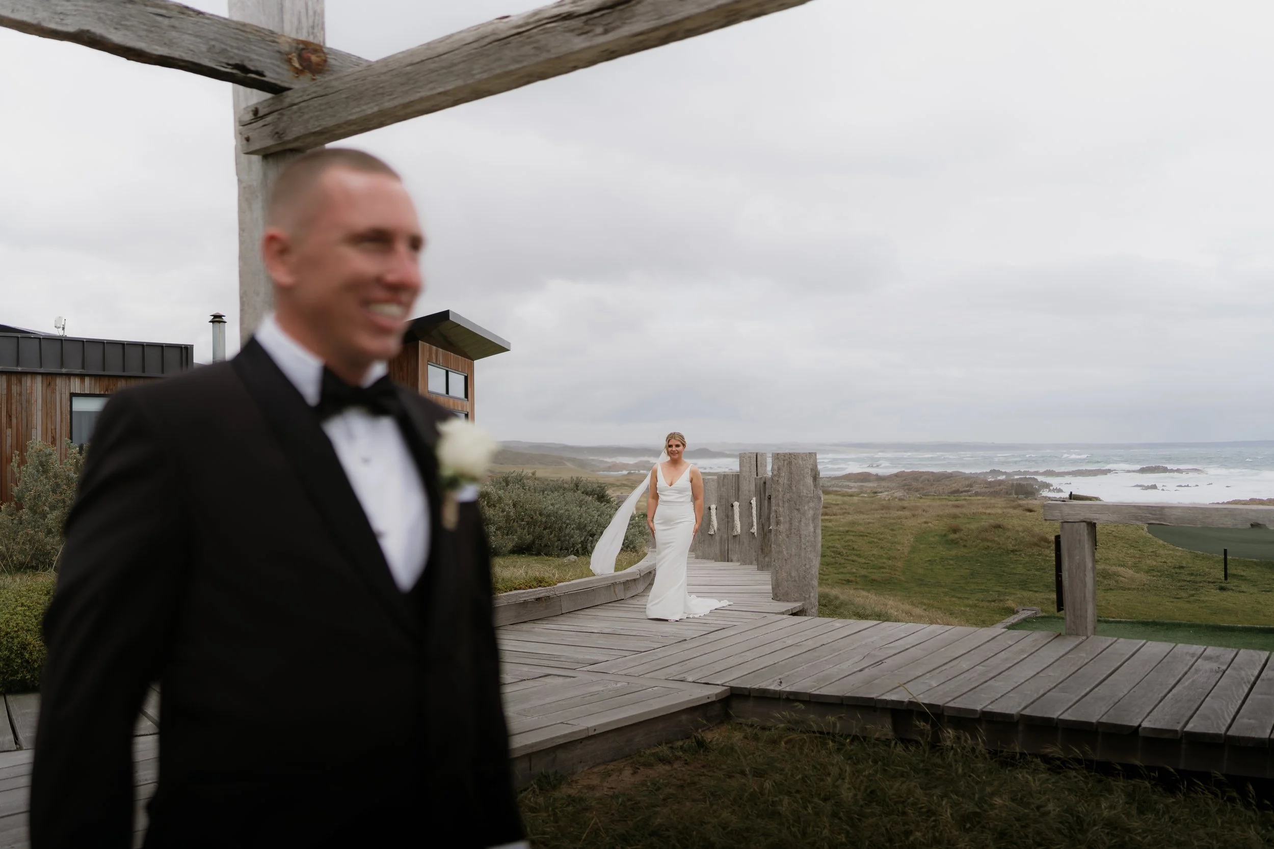 A man in a tuxedo smiling in the foreground, with a woman in a wedding dress standing on a wooden walkway near the coast in the background.