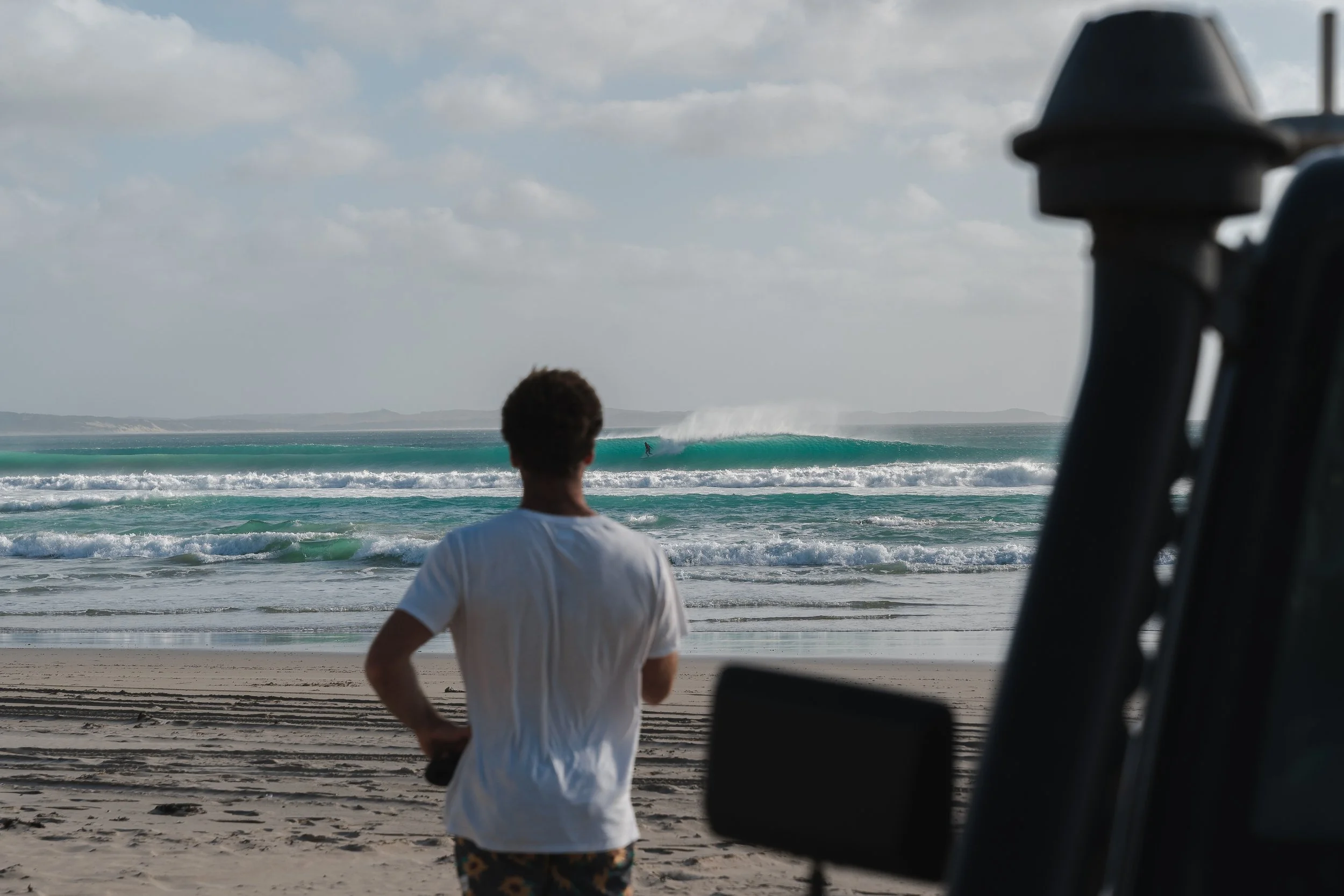 A person wearing a white shirt and shorts with a colorful pattern standing on a sandy beach, looking at the ocean where a surfer is riding a wave, seen through the window of a vehicle.