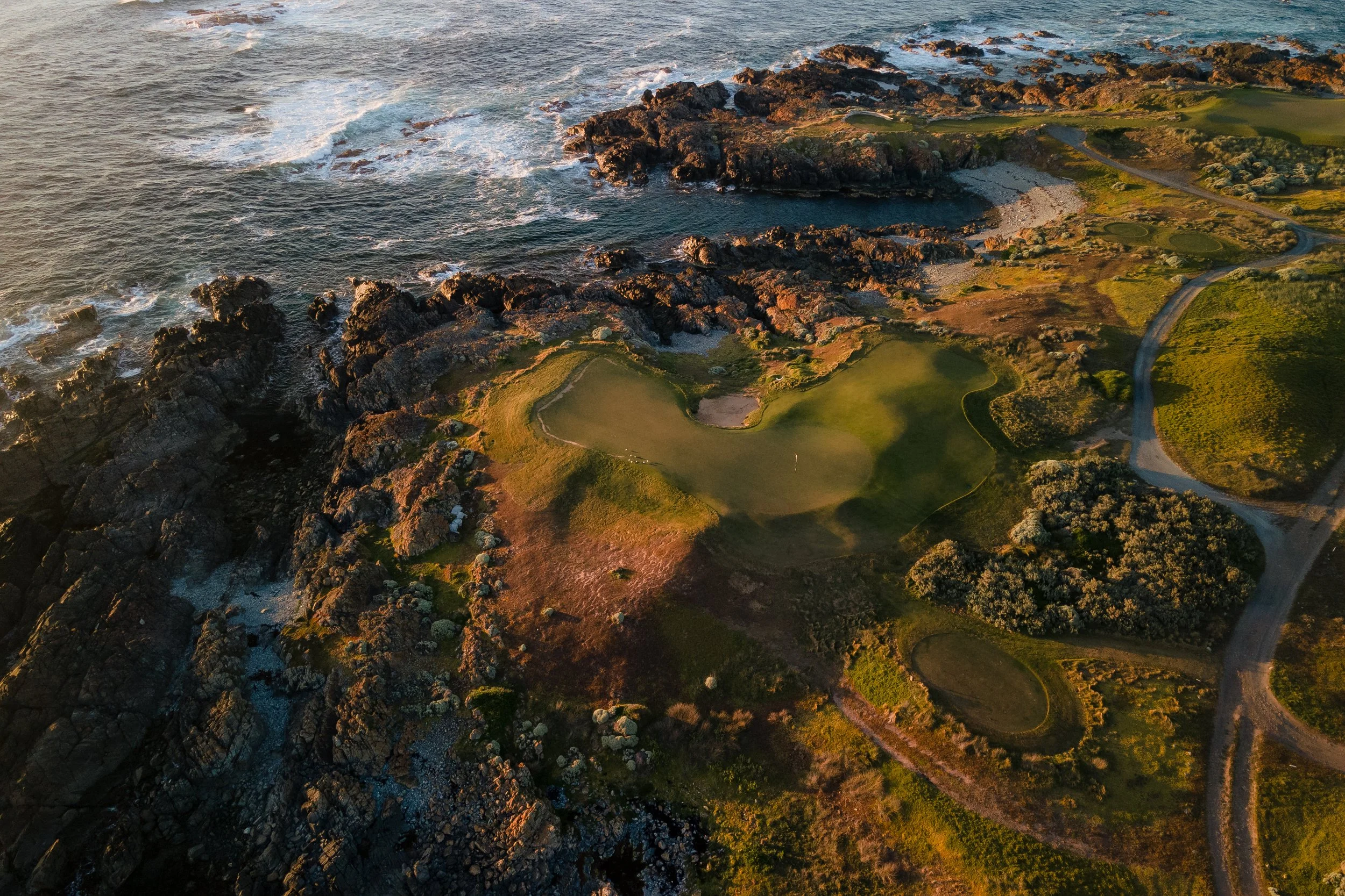 Aerial view of a coastline with rocky formations, sandy beach, and grassy areas, with ocean waves crashing onto the rocks.