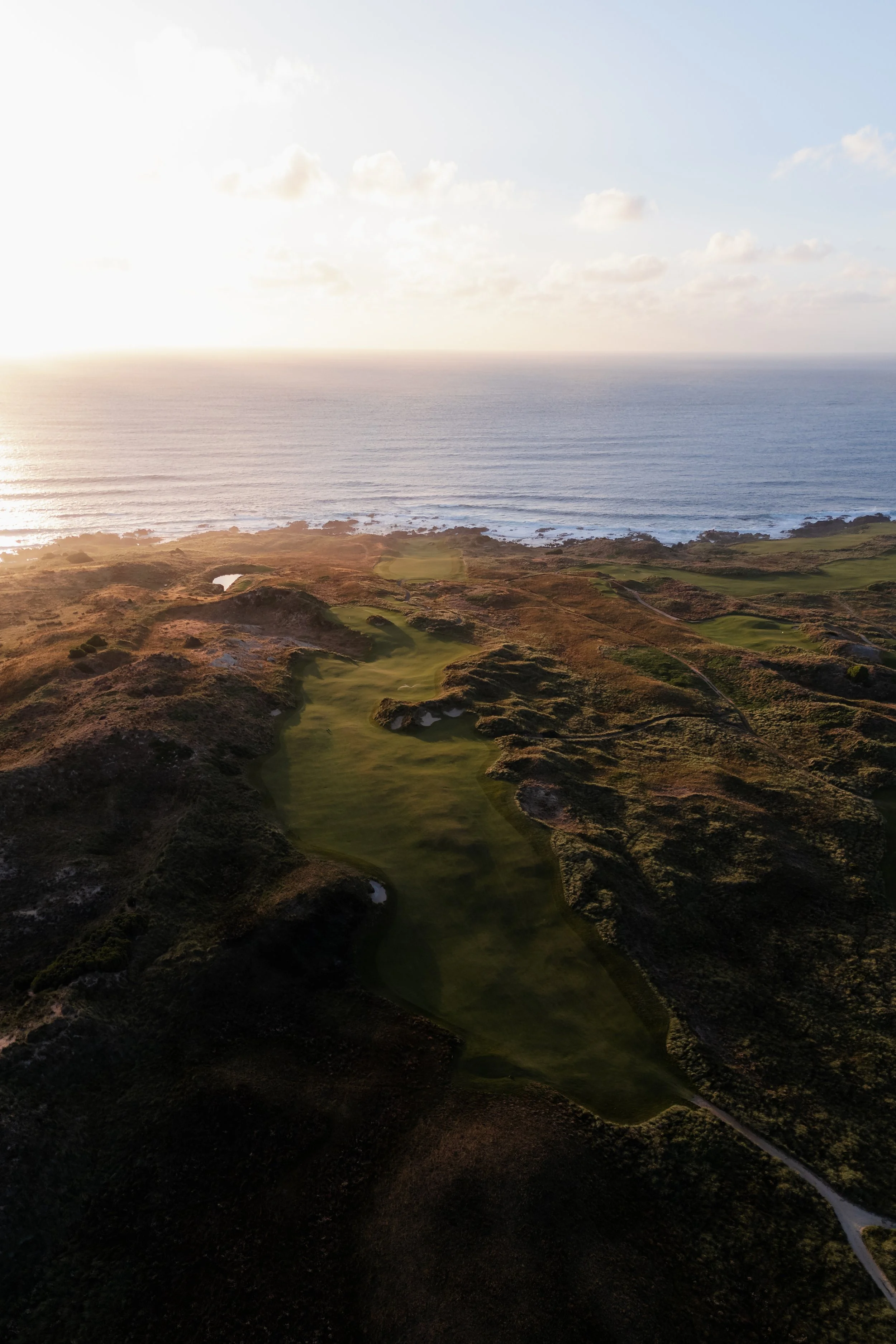 View of a coastal golf course at sunset, with the ocean in the background and a winding path through the course.